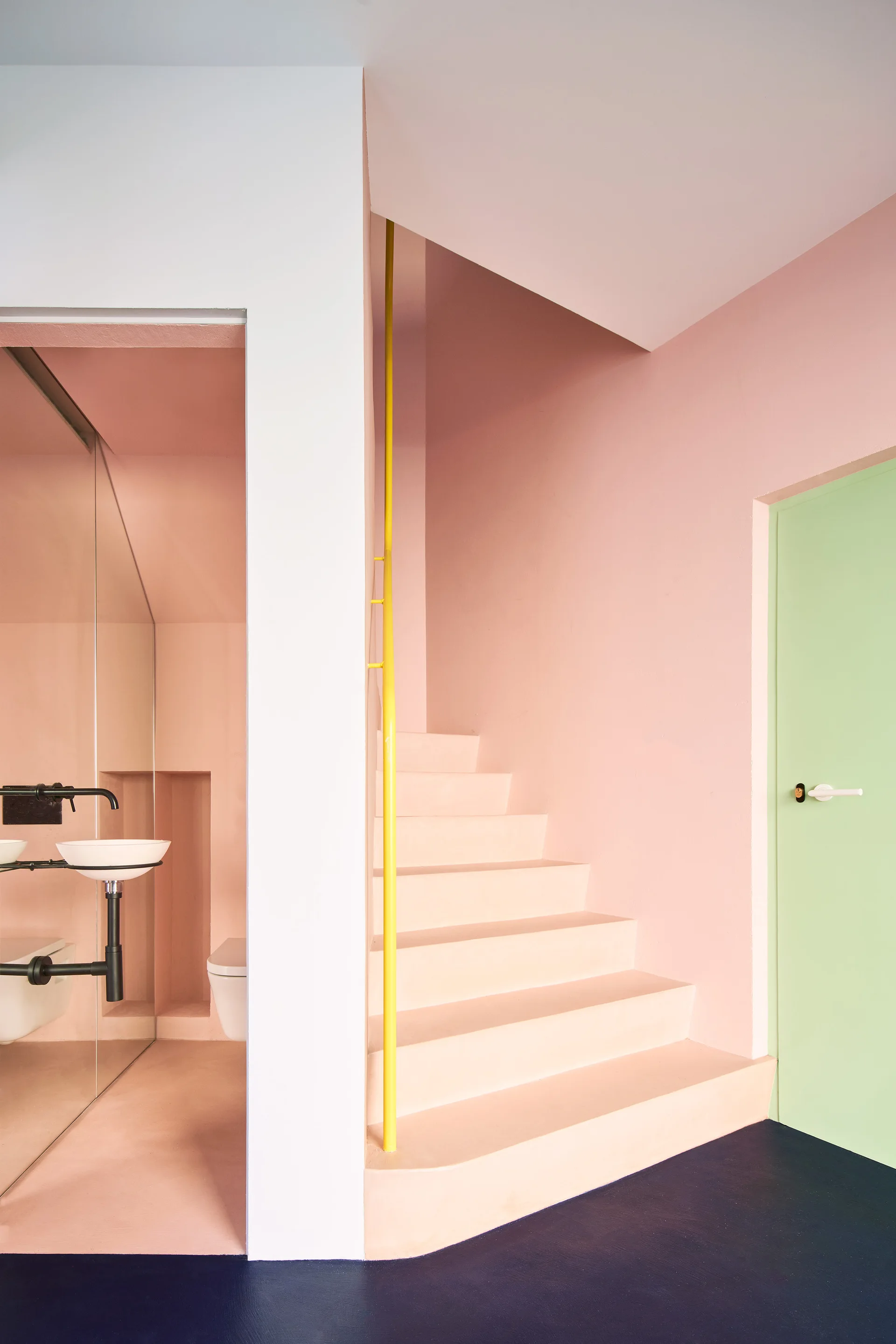 A pink minimalist staircase features a bright yellow vertical handrail next to a light green door and a glass-partitioned bathroom.