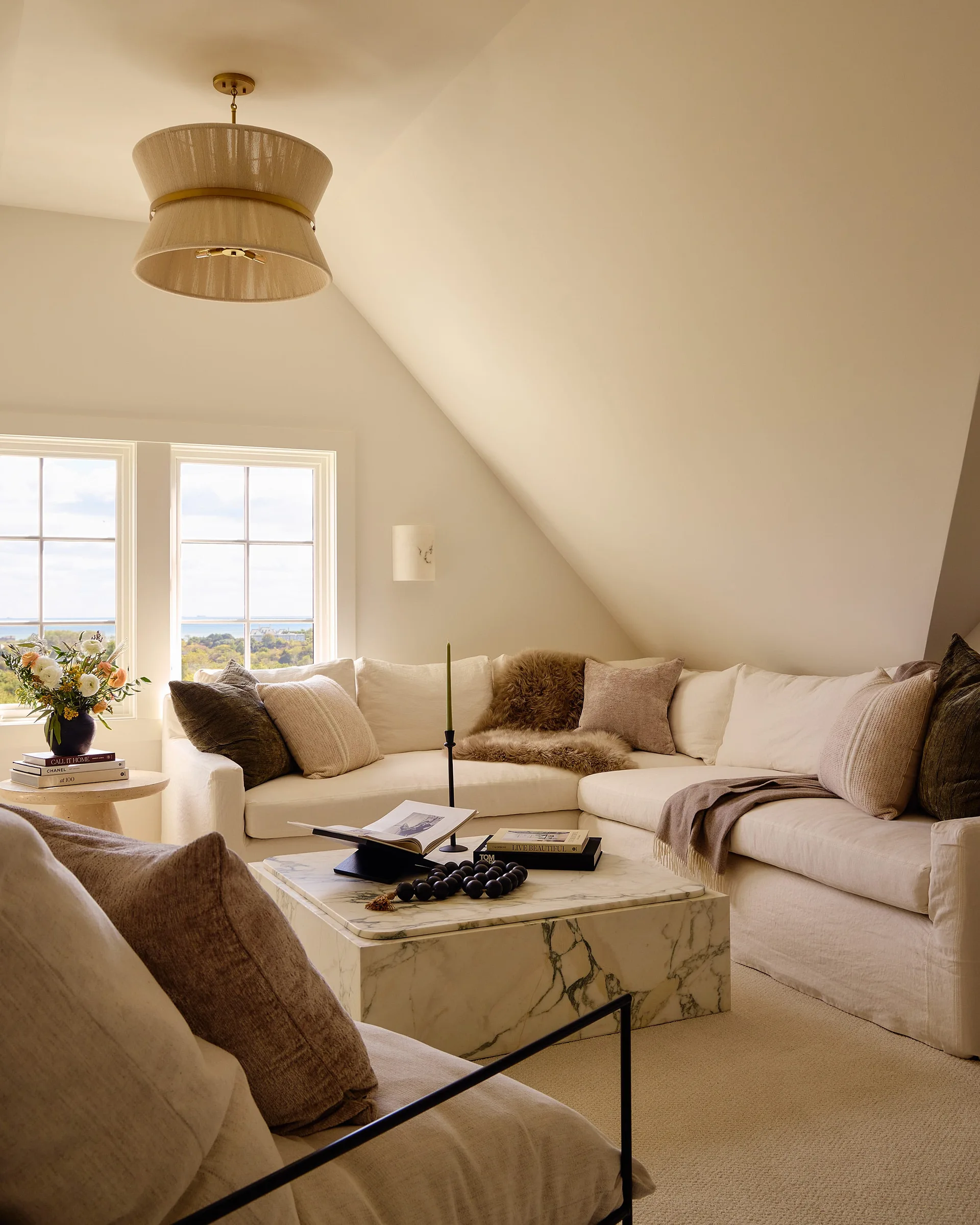 A cozy attic living room features a cream sectional sofa, a low square marble coffee table, and a fabric drum pendant light under a sloped ceiling.