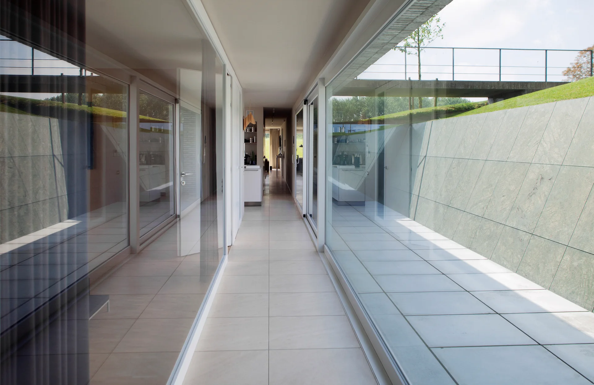 A long, minimalist hallway featuring floor-to-ceiling glass walls, light tiled floors, and views of an exterior stone retaining wall.