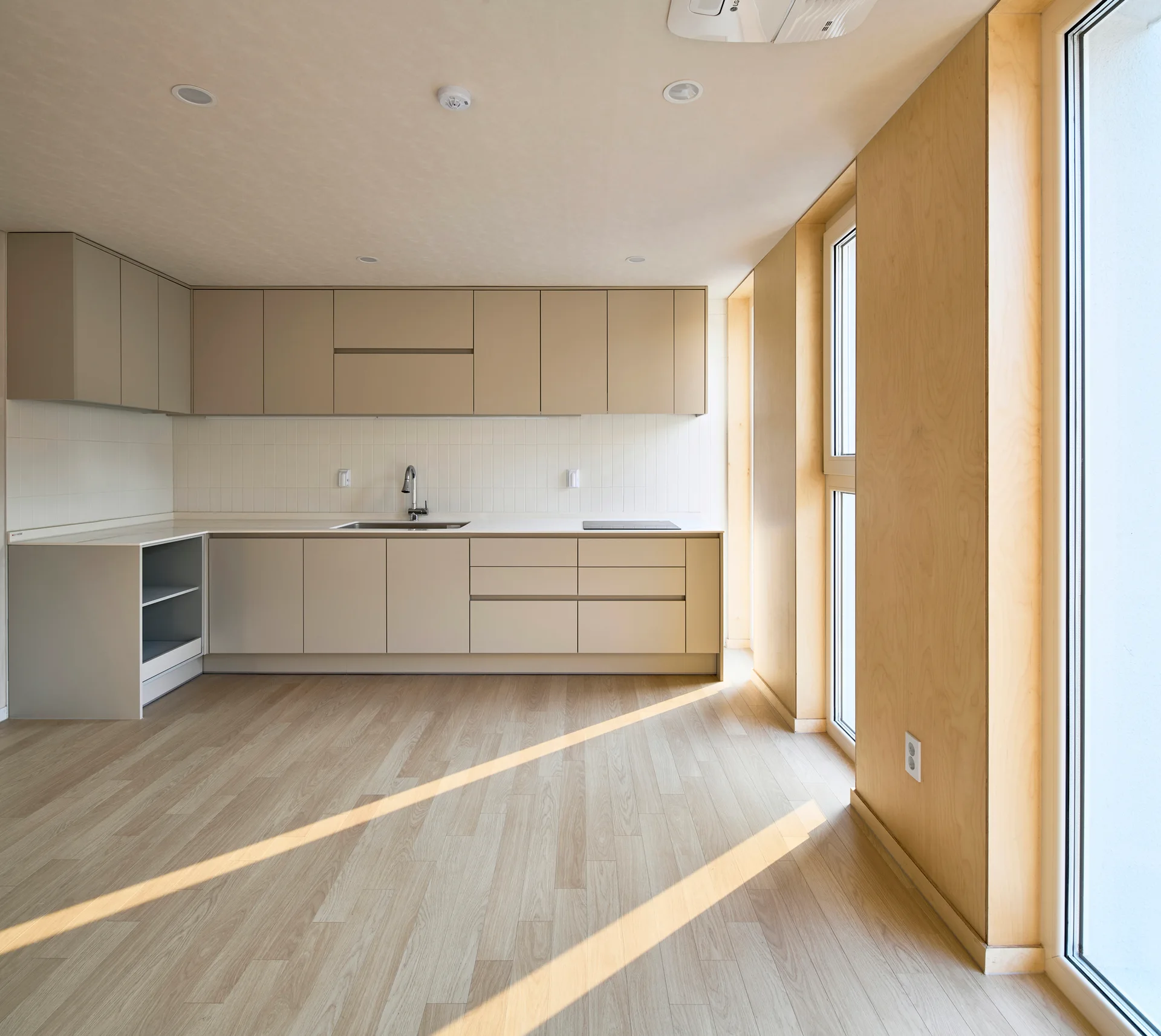 A minimalist kitchen features beige flat-panel cabinetry, a vertical white tiled backsplash, and light wood flooring under natural sunlight.