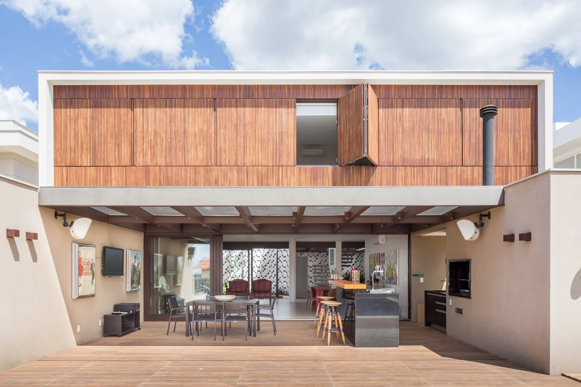 A covered outdoor patio featuring a wooden dining set, a stone-topped bar with stools, and a vertical wood slat facade on the second floor.