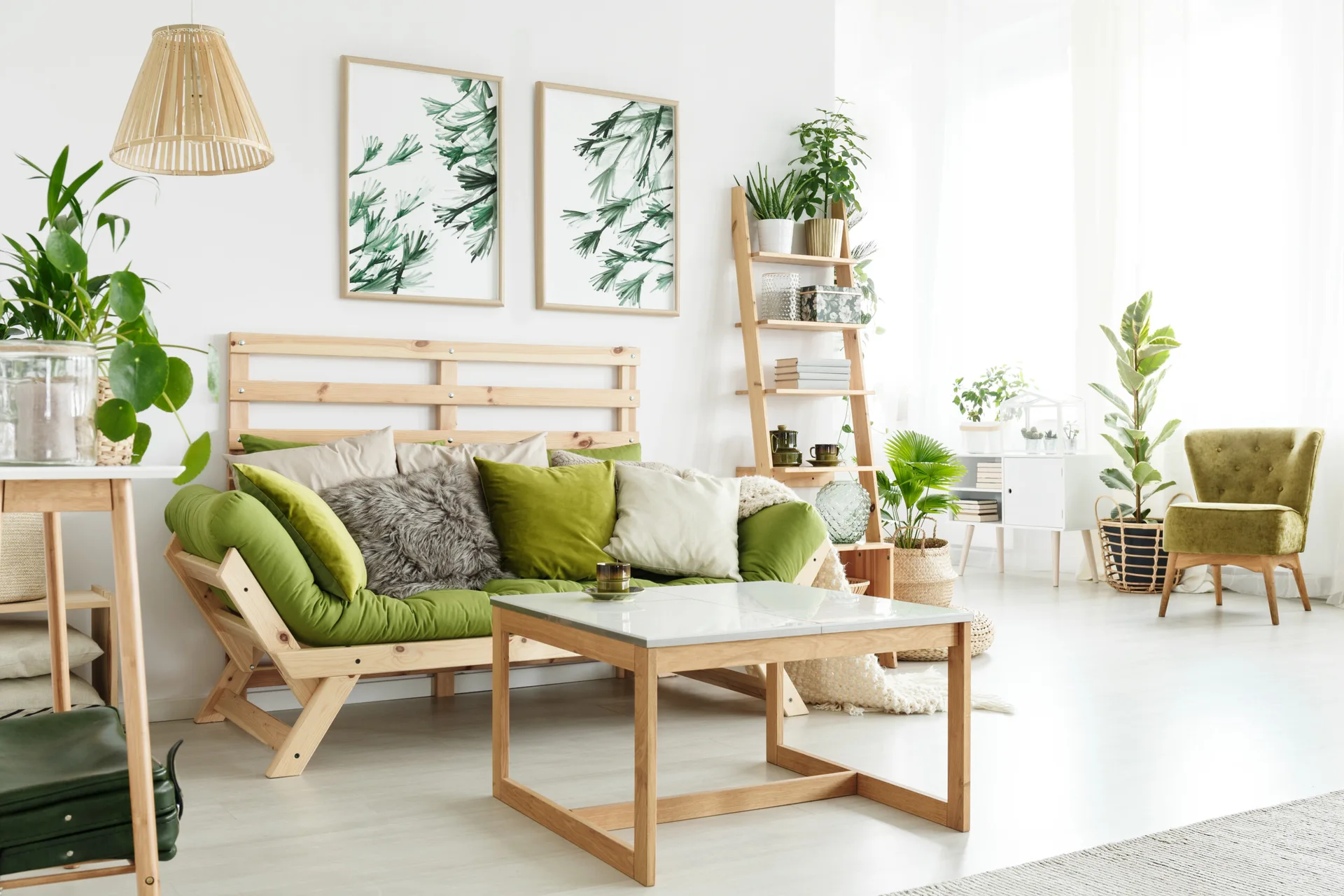 A bright Scandinavian living room featuring a green wooden futon, a coffee table, a ladder shelf with plants, and botanical framed prints.
