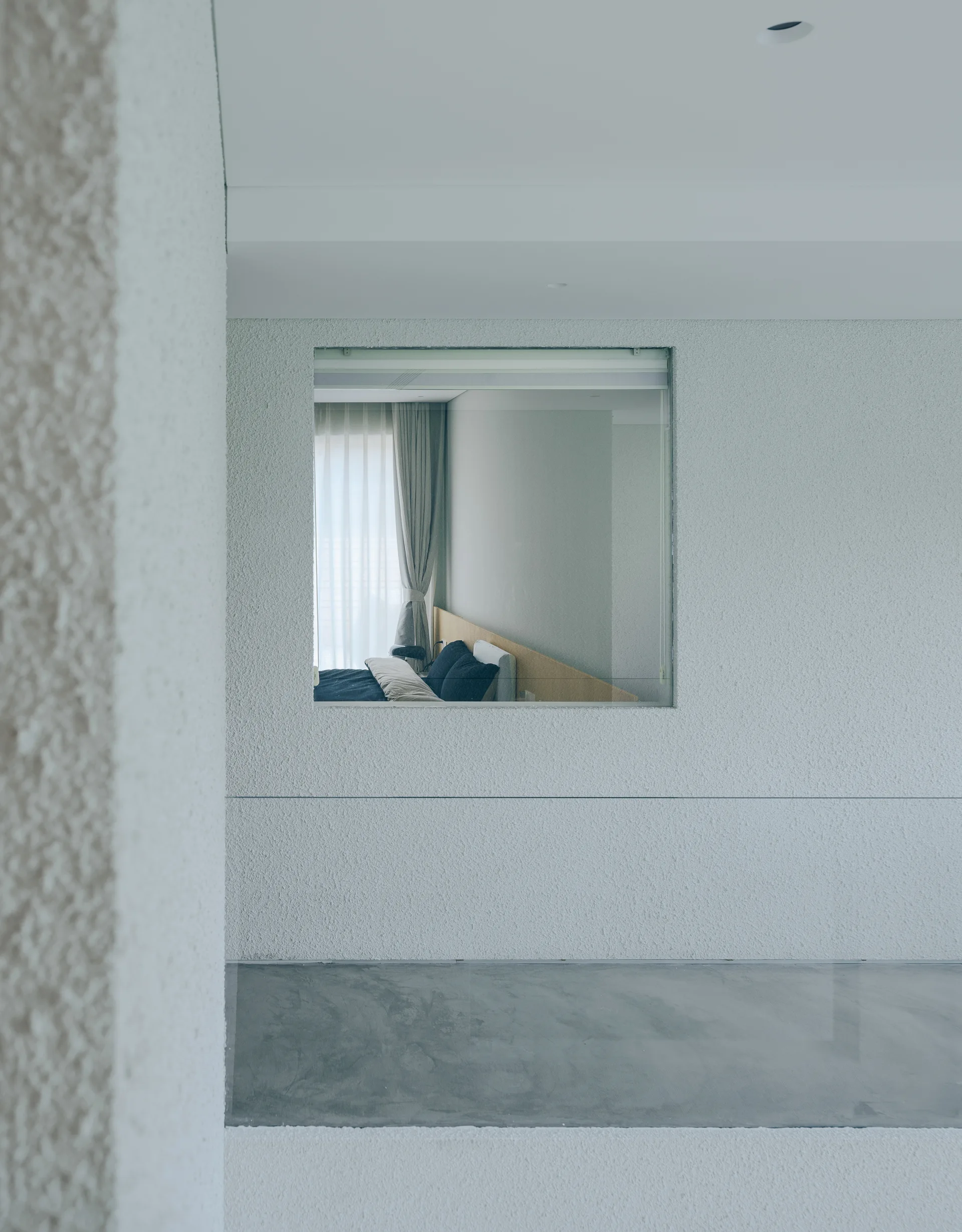 A view through a square interior wall opening showing a bedroom with a wood headboard, dark pillows, and neutral drapery.