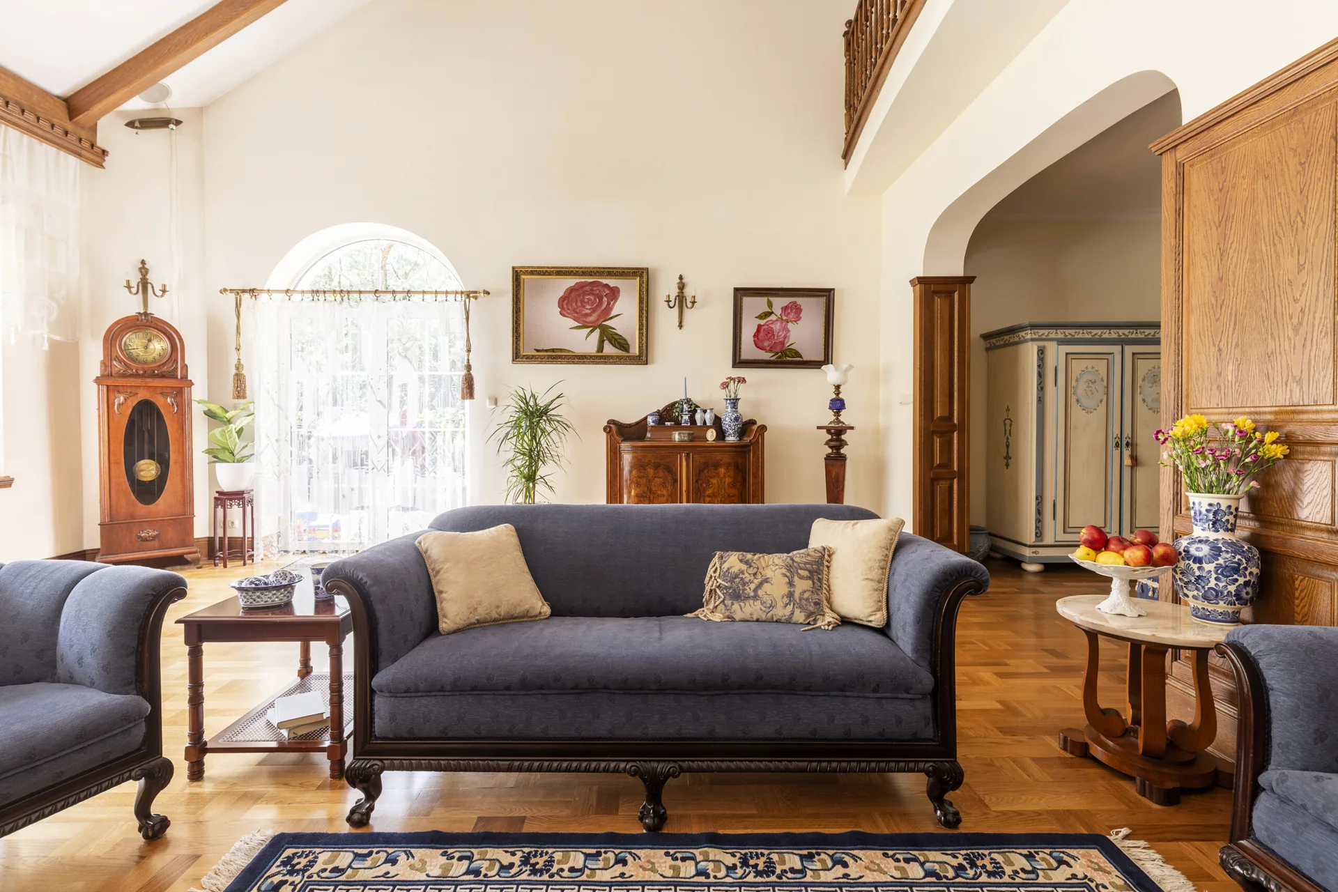 A traditional living room features blue upholstered sofas, polished parquet wood flooring, a grandfather clock, and cream walls under a vaulted ceiling.