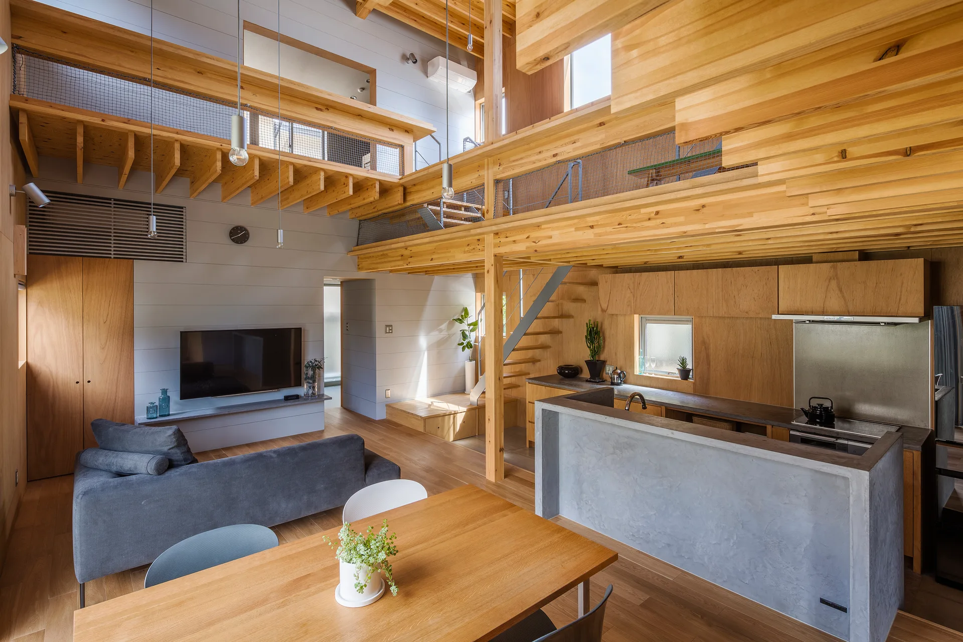A double-height residential living area features extensive light wood beams, a mezzanine with net railings, a grey sofa, and a concrete kitchen island.