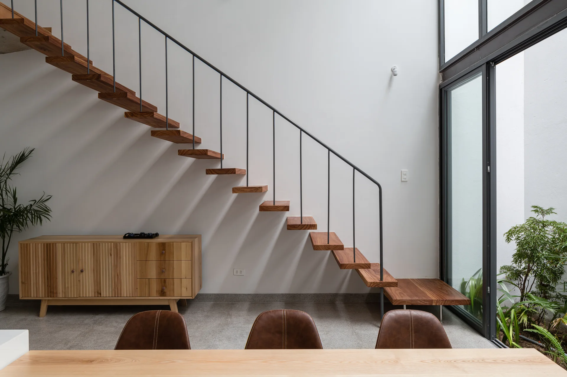 Modern floating wood stairs with a black metal railing rise above a wooden credenza and next to a glass wall with plants.