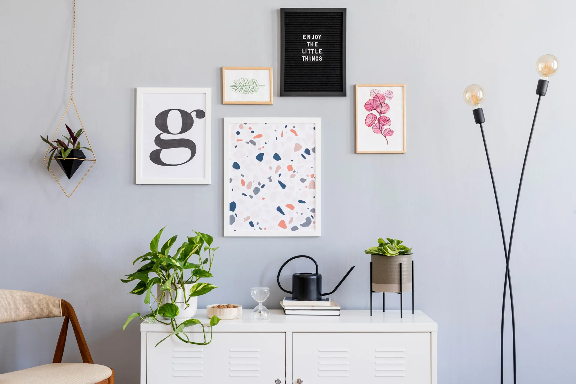 A white metal locker-style cabinet sits against a light grey wall decorated with a gallery wall of framed art, potted plants, and a slim floor lamp.