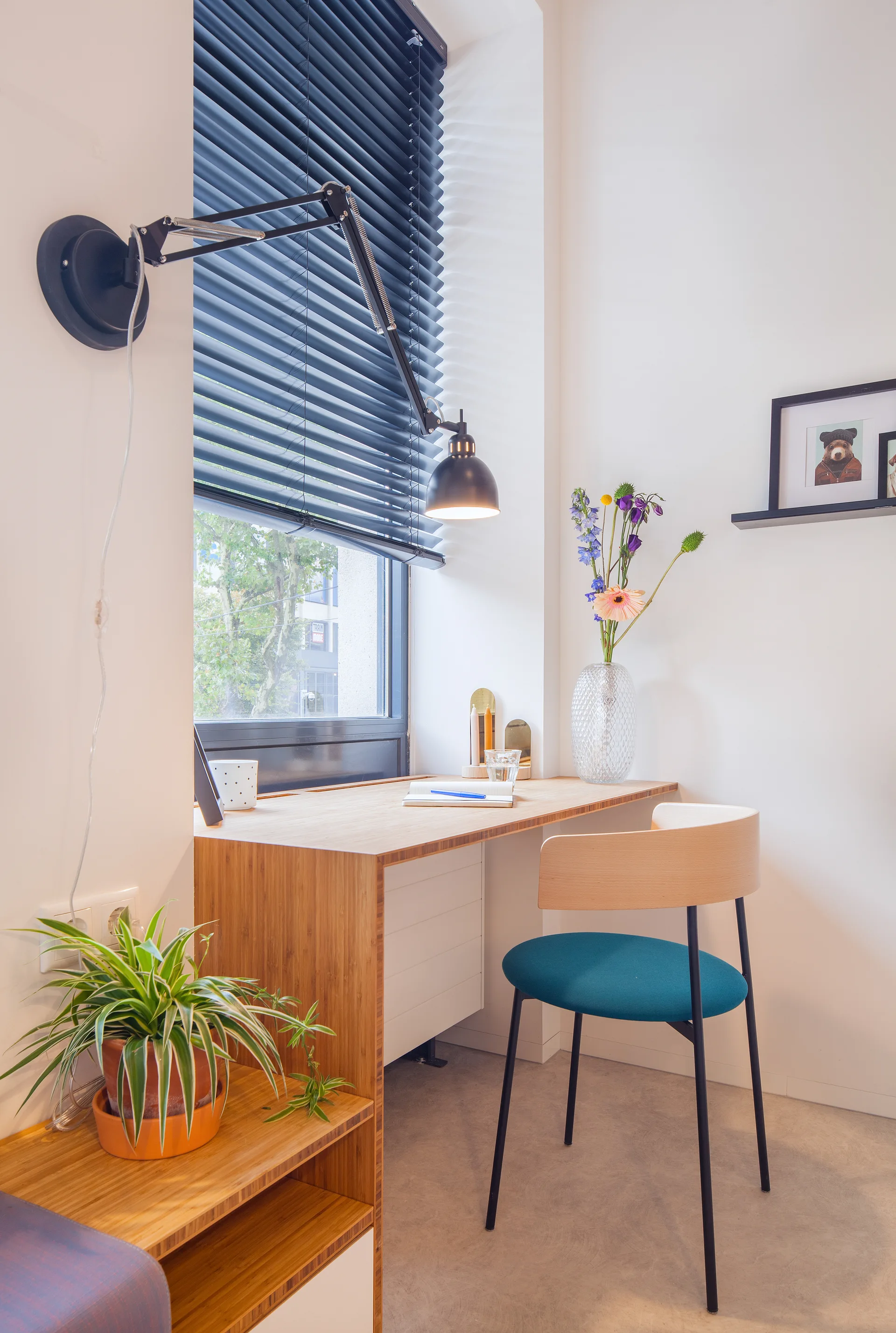 A small home office corner features a wooden desk in a window nook, a chair with a blue cushion, and a black wall-mounted lamp.