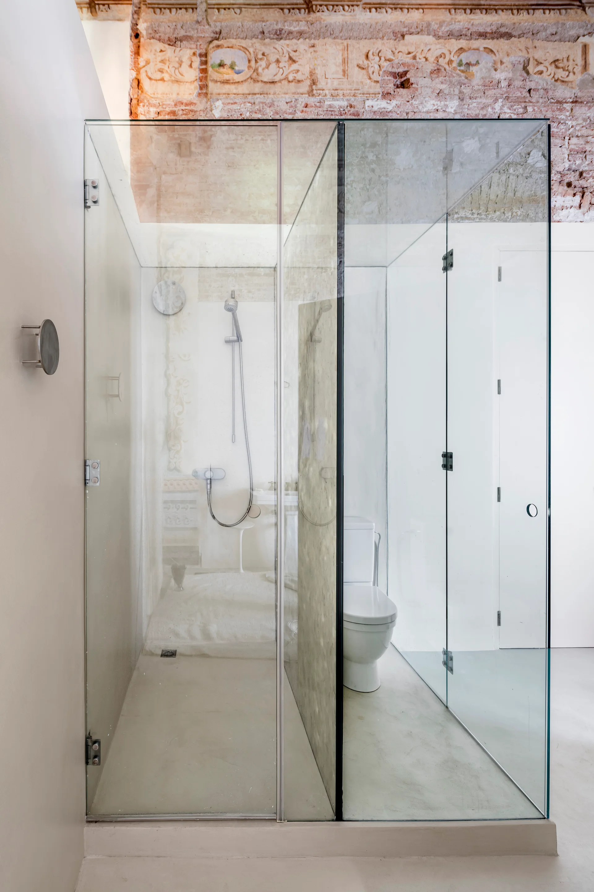 A glass shower enclosure stands beneath an exposed brick and plaster ceiling with a faded historic mural in a minimalist bathroom.