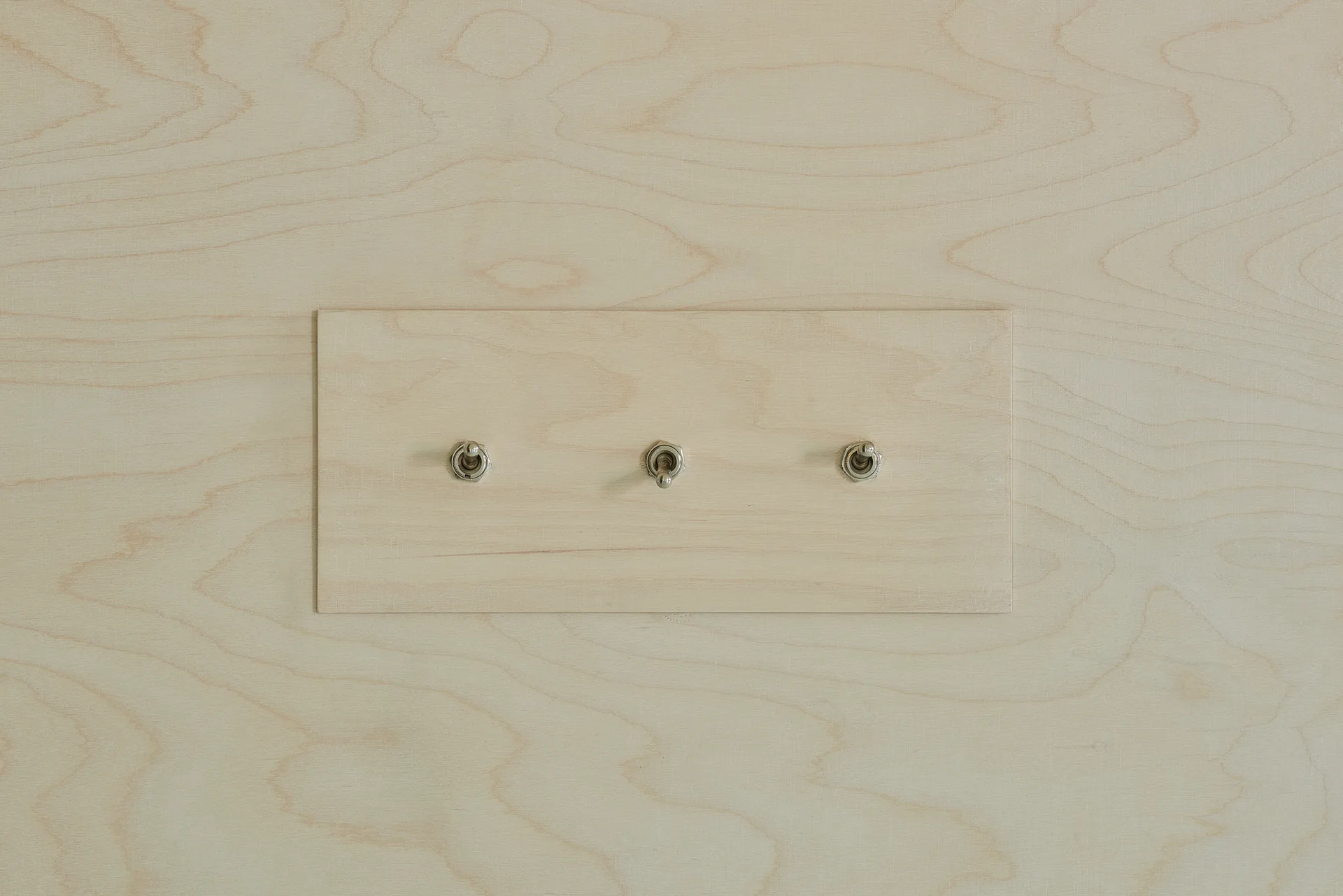 Three silver metal toggle switches mounted on a matching rectangular plywood plate integrated into a light wood grain plywood wall.