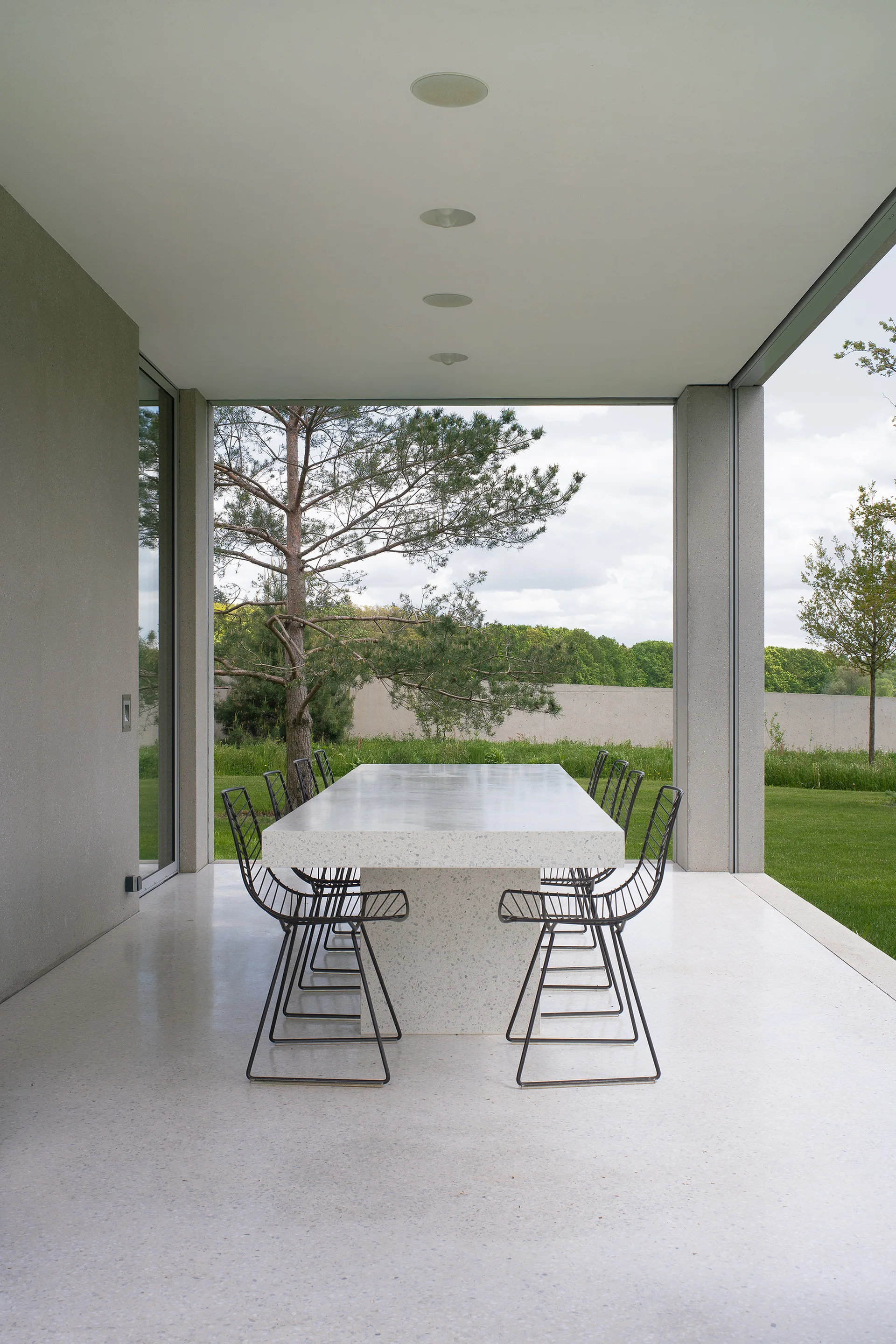 A modern covered concrete patio features a large speckled terrazzo dining table paired with black wire chairs looking out onto a grassy lawn.