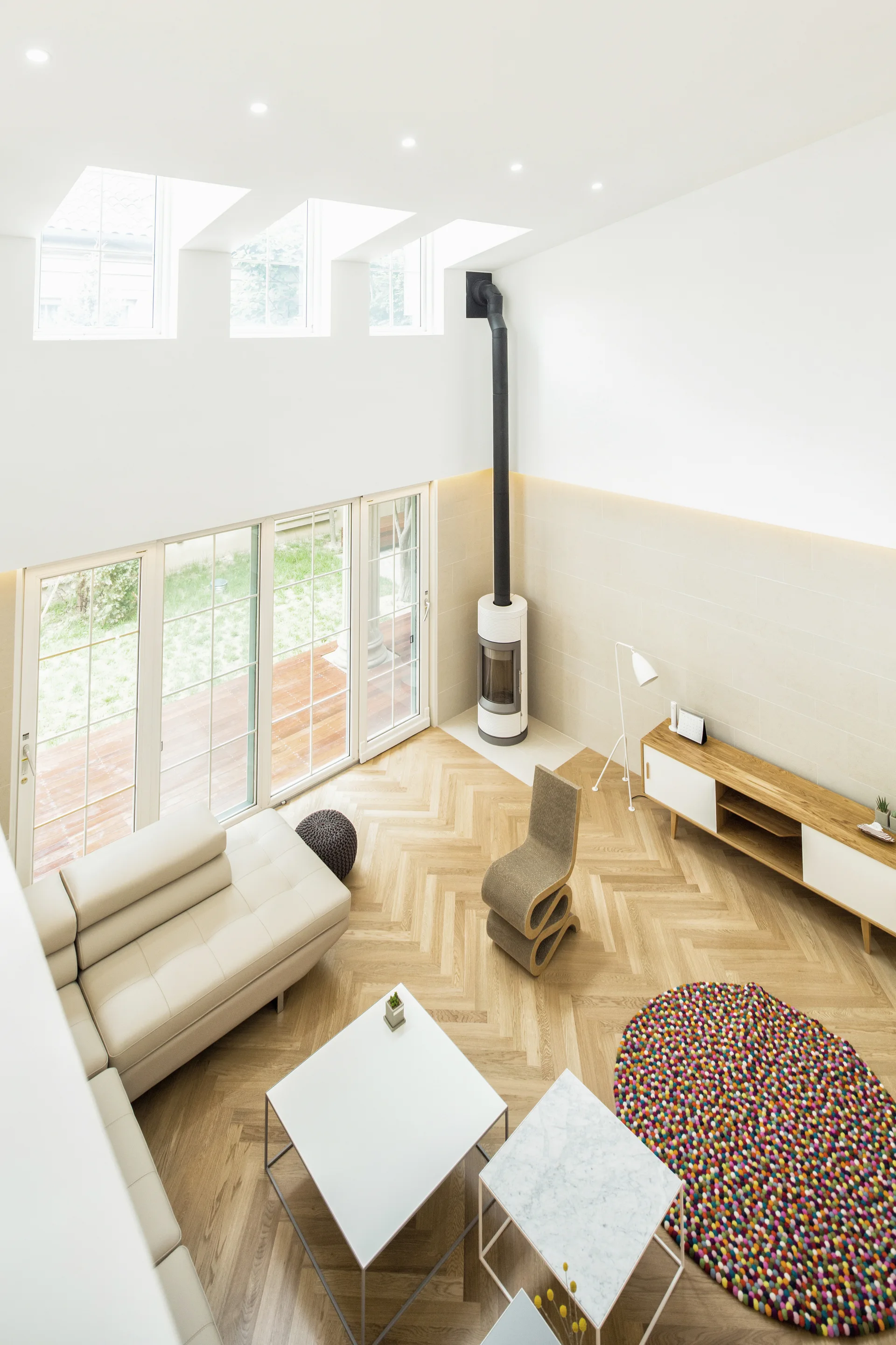 A high-angle view of a Scandinavian living room featuring light herringbone wood floors, a white sectional sofa, and a modern cylindrical stove.