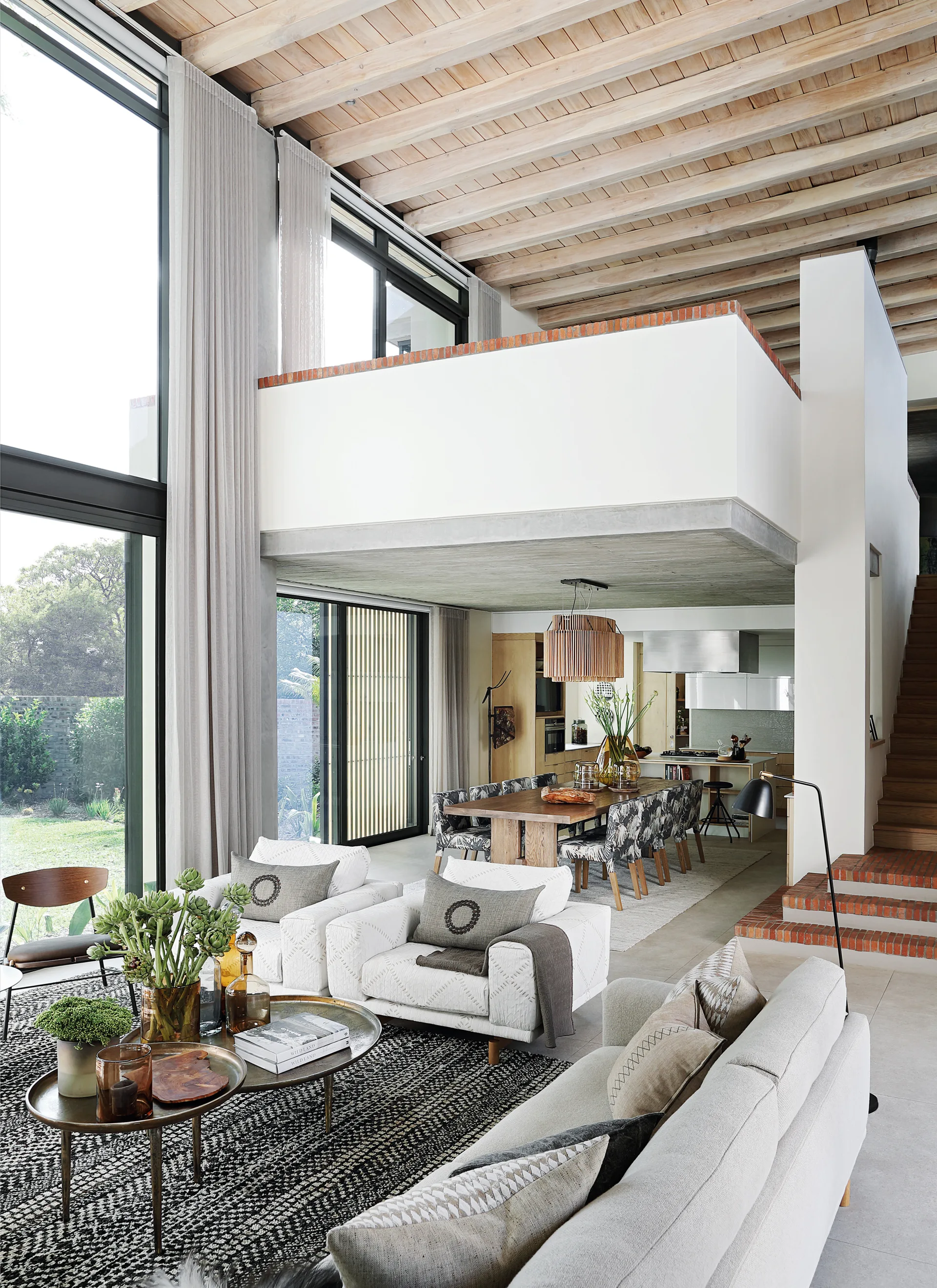 A double-height living room featuring exposed wood ceiling beams, white armchairs, a patterned rug, and a dining area under a mezzanine balcony.