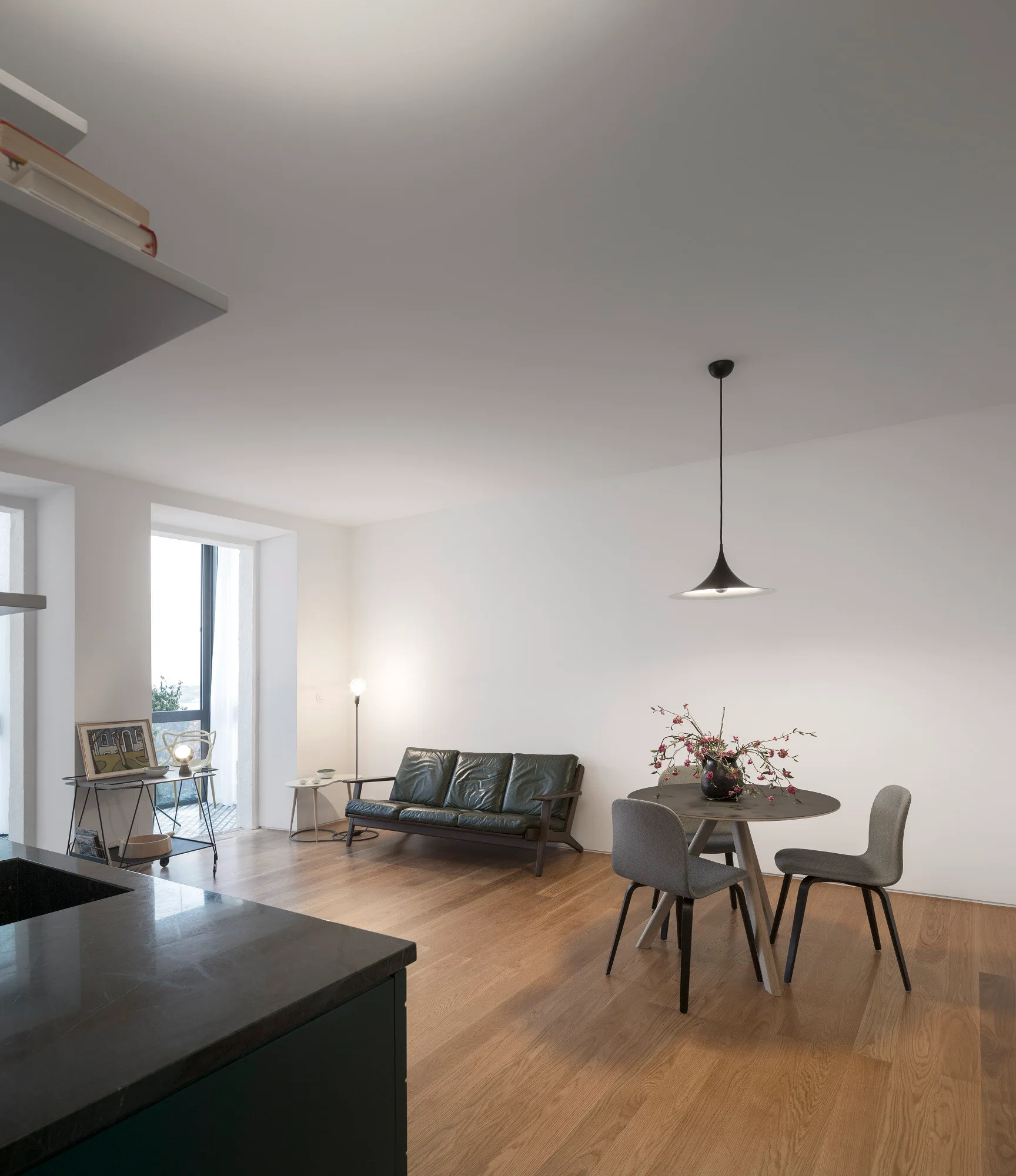 A minimalist open-plan room features a dark green leather sofa, a round wooden dining table with grey chairs, and a black pendant light on wood floors.