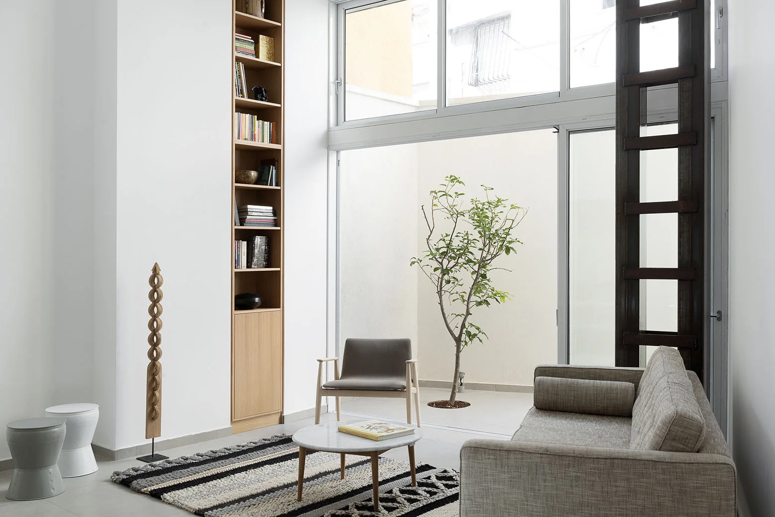 A light gray sofa, wooden armchair, and marble coffee table sit near a tall wood bookshelf and a glass door opening to a courtyard with a small tree.