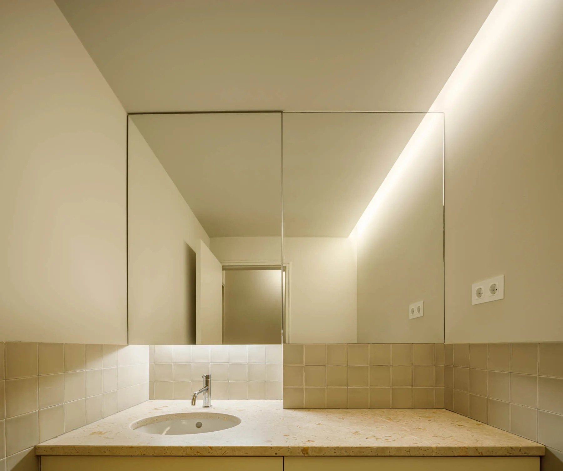 A minimalist bathroom vanity features a stone countertop, a square-tiled backsplash, a mirrored cabinet, and warm overhead cove lighting.