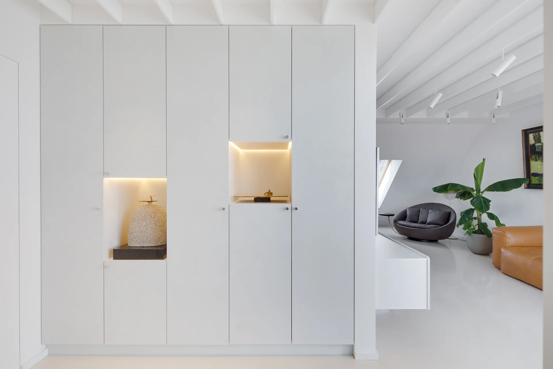 Matte white floor-to-ceiling cabinets feature two illuminated display niches displaying sculptures next to a bright modern living room.