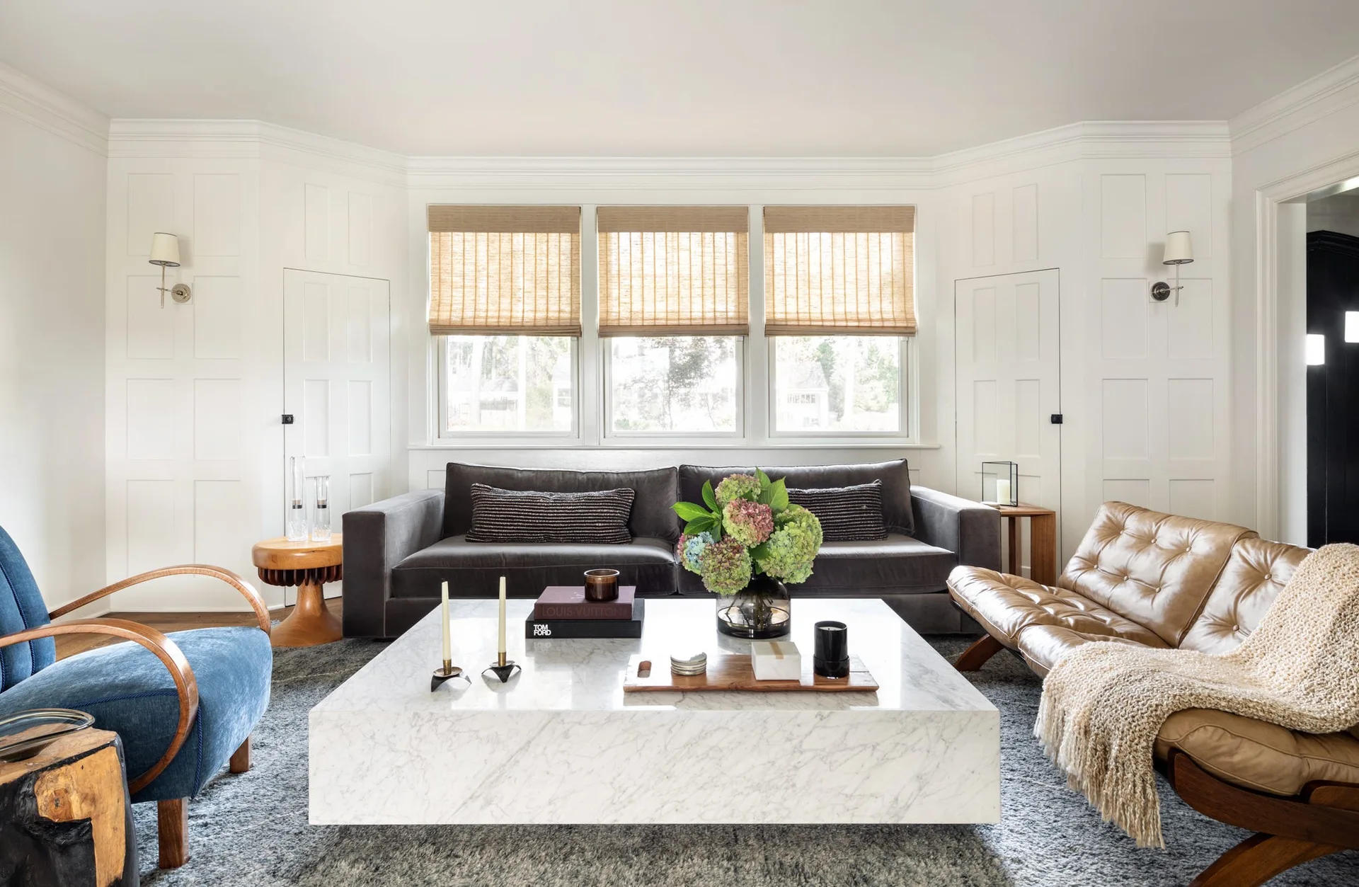 A living room featuring a large square white marble coffee table, a grey velvet sofa, a tan leather chair, and three woven window shades.