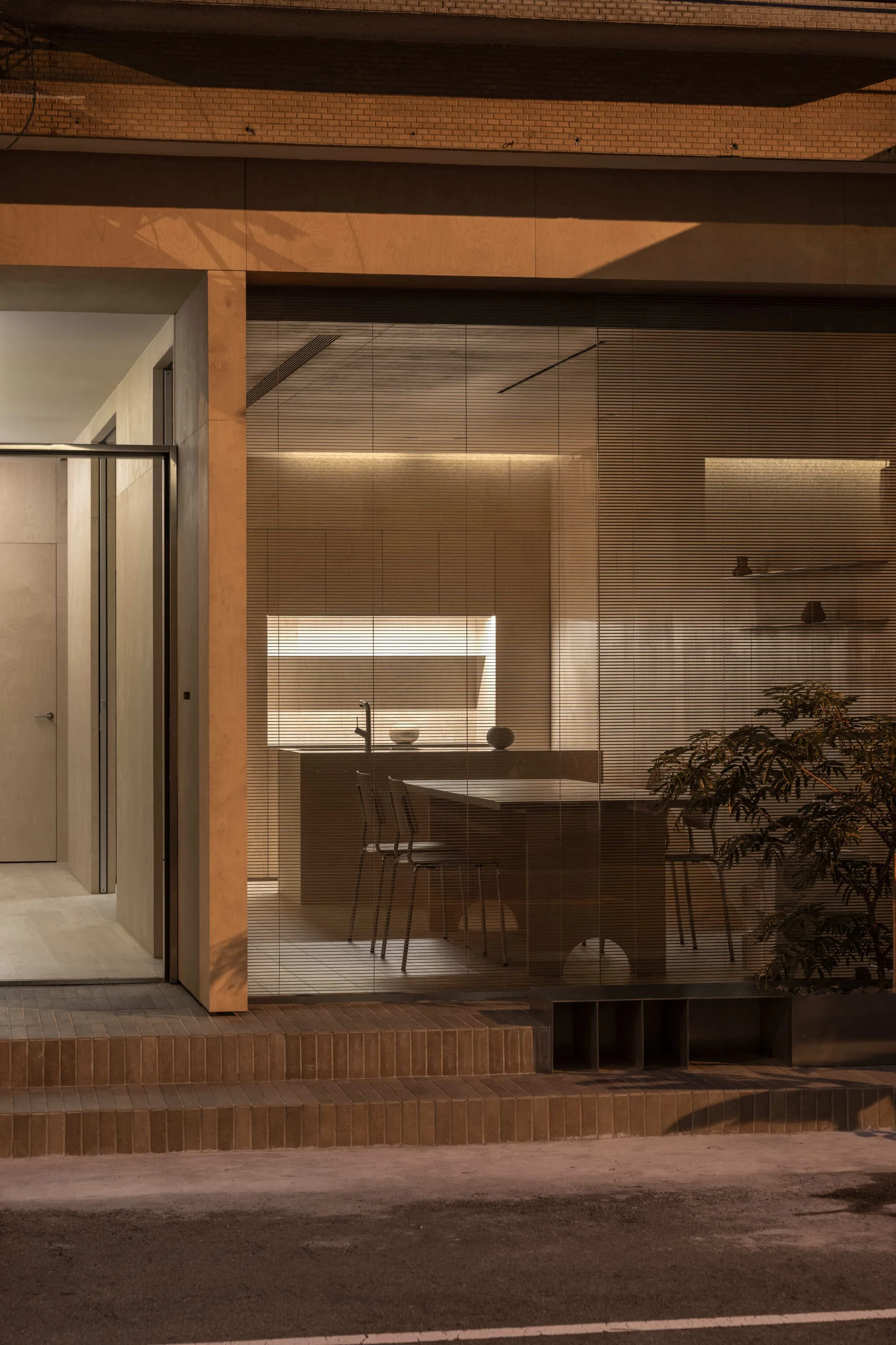 A minimalist kitchen with warm plywood walls and a central island is visible through a large glass facade with horizontal blinds.