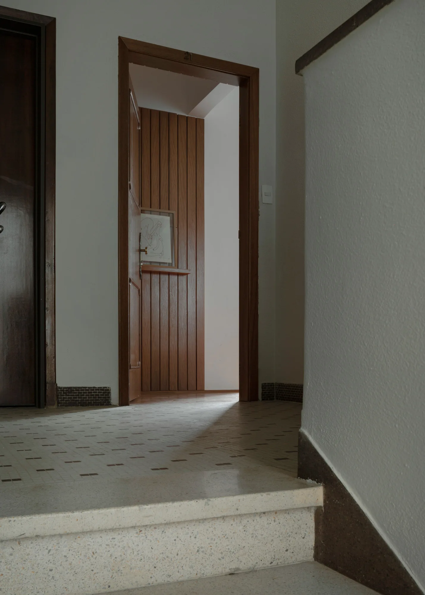 A view from terrazzo stairs looking onto a tiled hallway landing with wood door frames and an open doorway showing vertical wood paneling.
