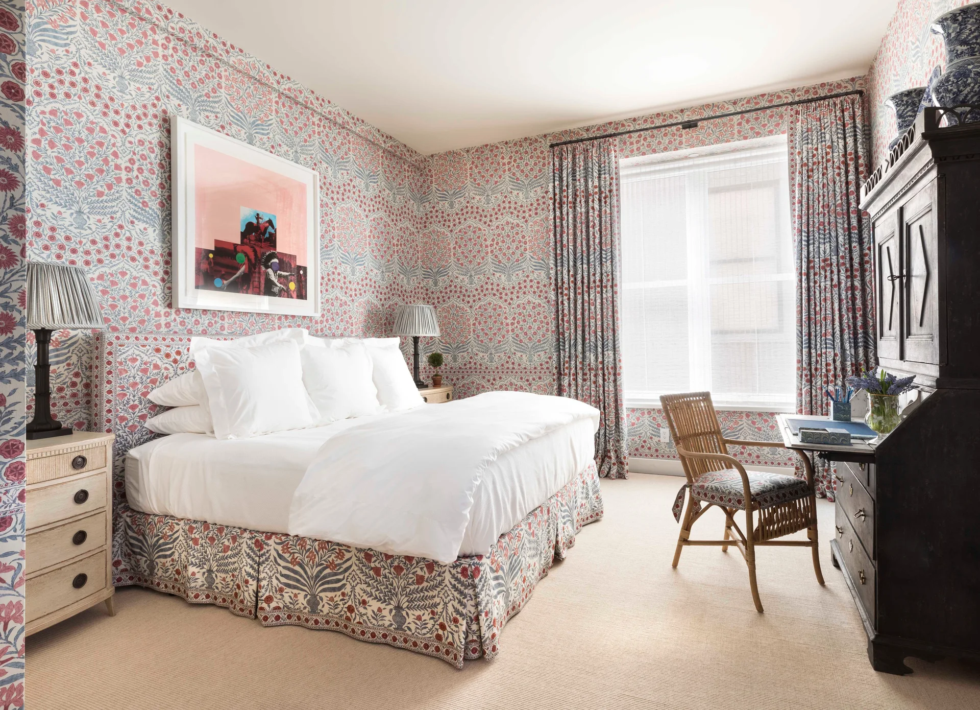 A traditional bedroom features matching floral wallpaper and curtains, a bed with white linens, and a dark wood secretary desk.