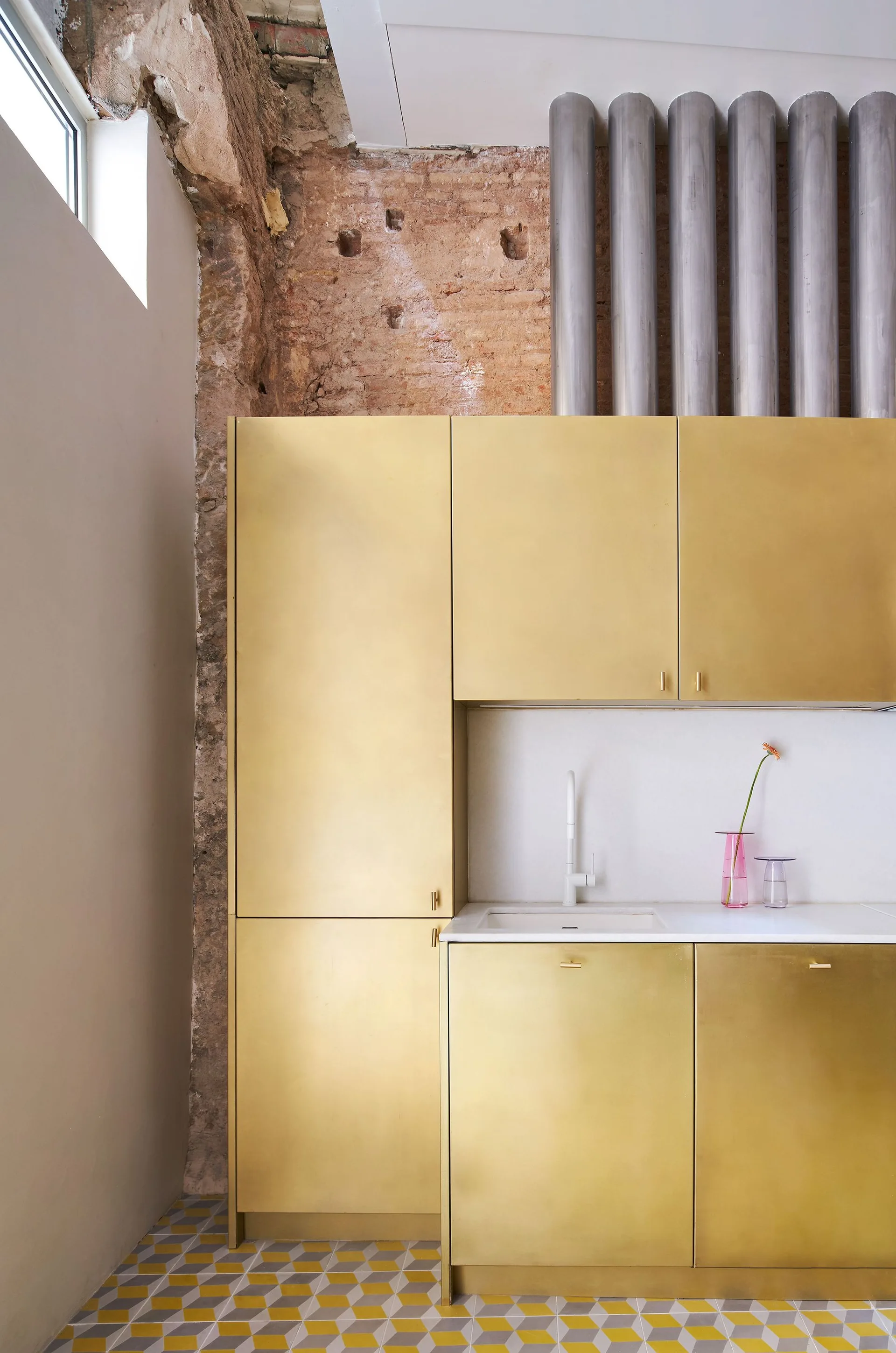 A modern kitchen corner featuring metallic brass cabinets, white solid countertop with sink, exposed brick, and grey and yellow geometric floor tiles.