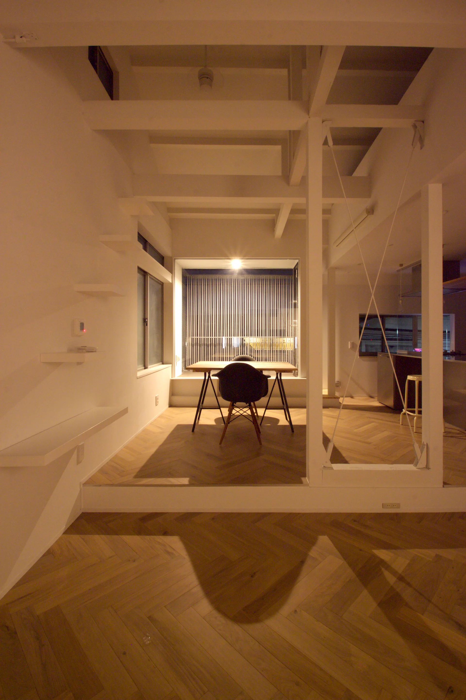 A modern minimalist interior featuring herringbone hardwood floors, a small dining table with a dark chair, and exposed white ceiling beams.