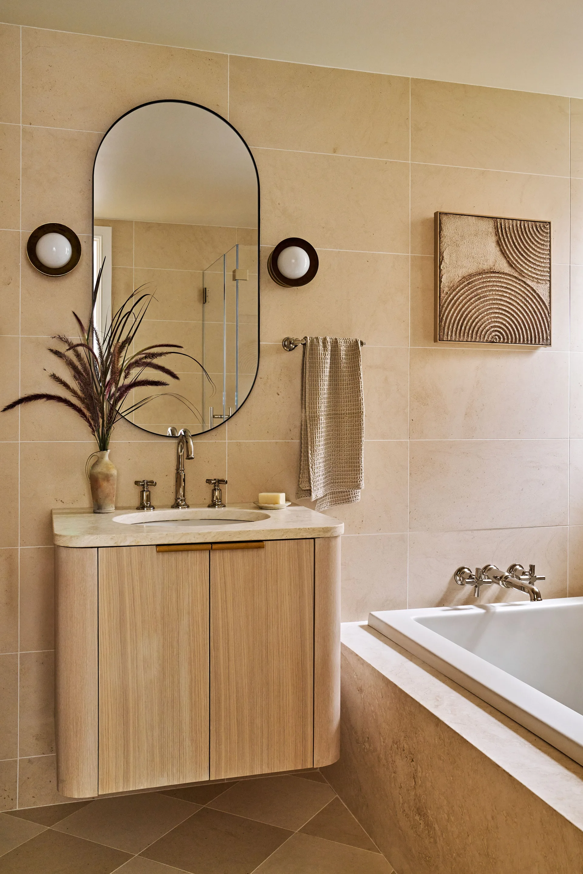 A modern bathroom showing a light wood floating vanity with a curved front, an oval black-framed mirror, two round sconces, and beige tiled walls.