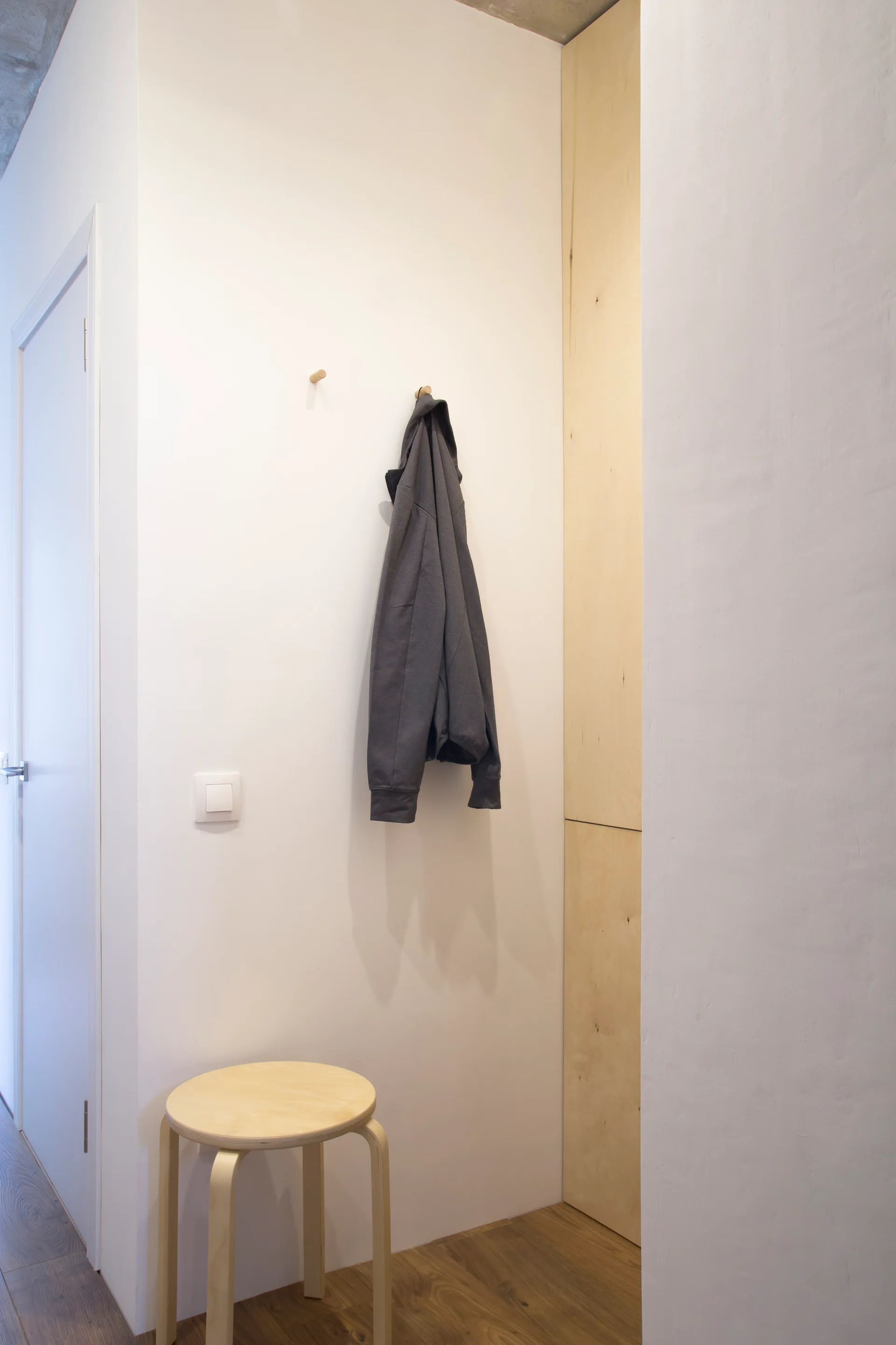 A minimalist entryway niche features white walls, a light wood built-in cabinet, wooden peg hooks, and a three-legged wooden stool.