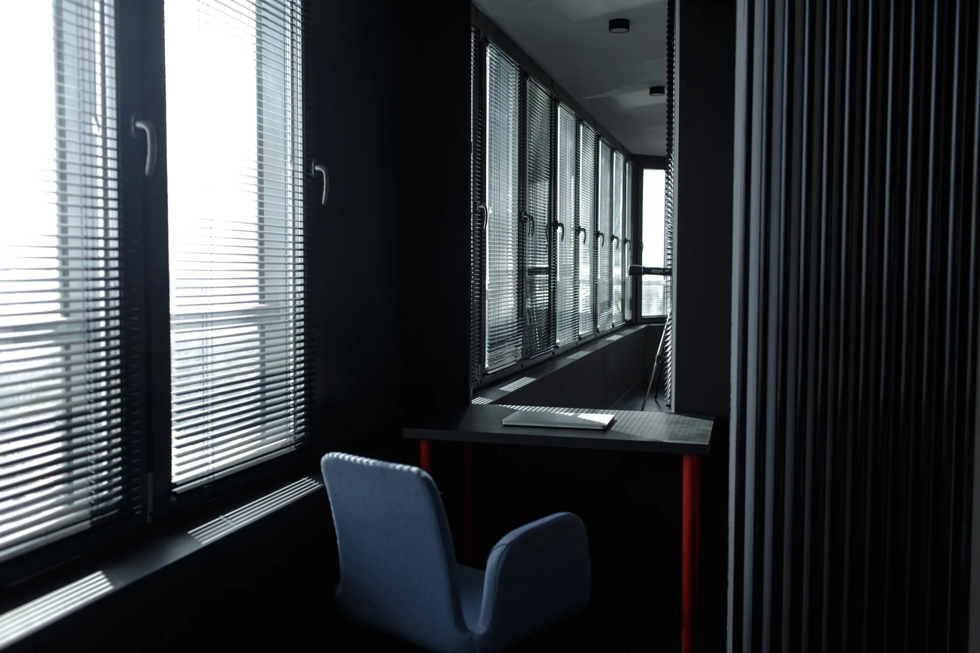 A dark minimalist home office features a desk with red legs, a grey upholstered chair, and large windows covered with horizontal venetian blinds.