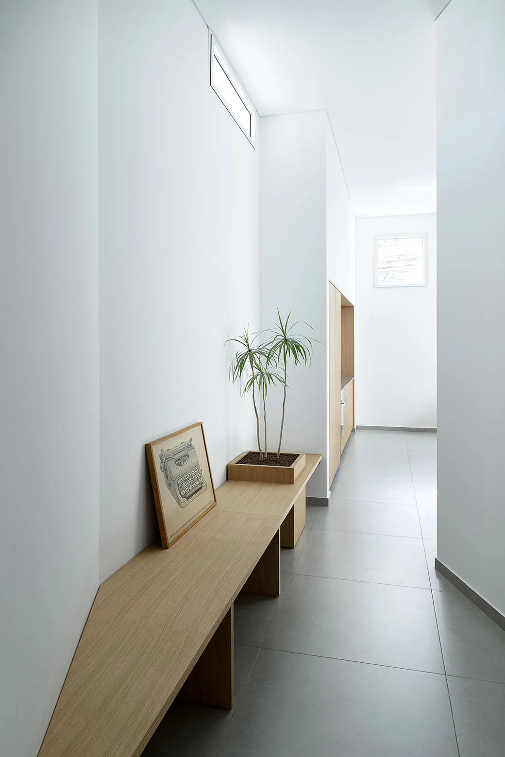 A long built-in wooden bench sits along a white minimalist hallway with grey tiled floors, a potted plant, and a high clerestory window.