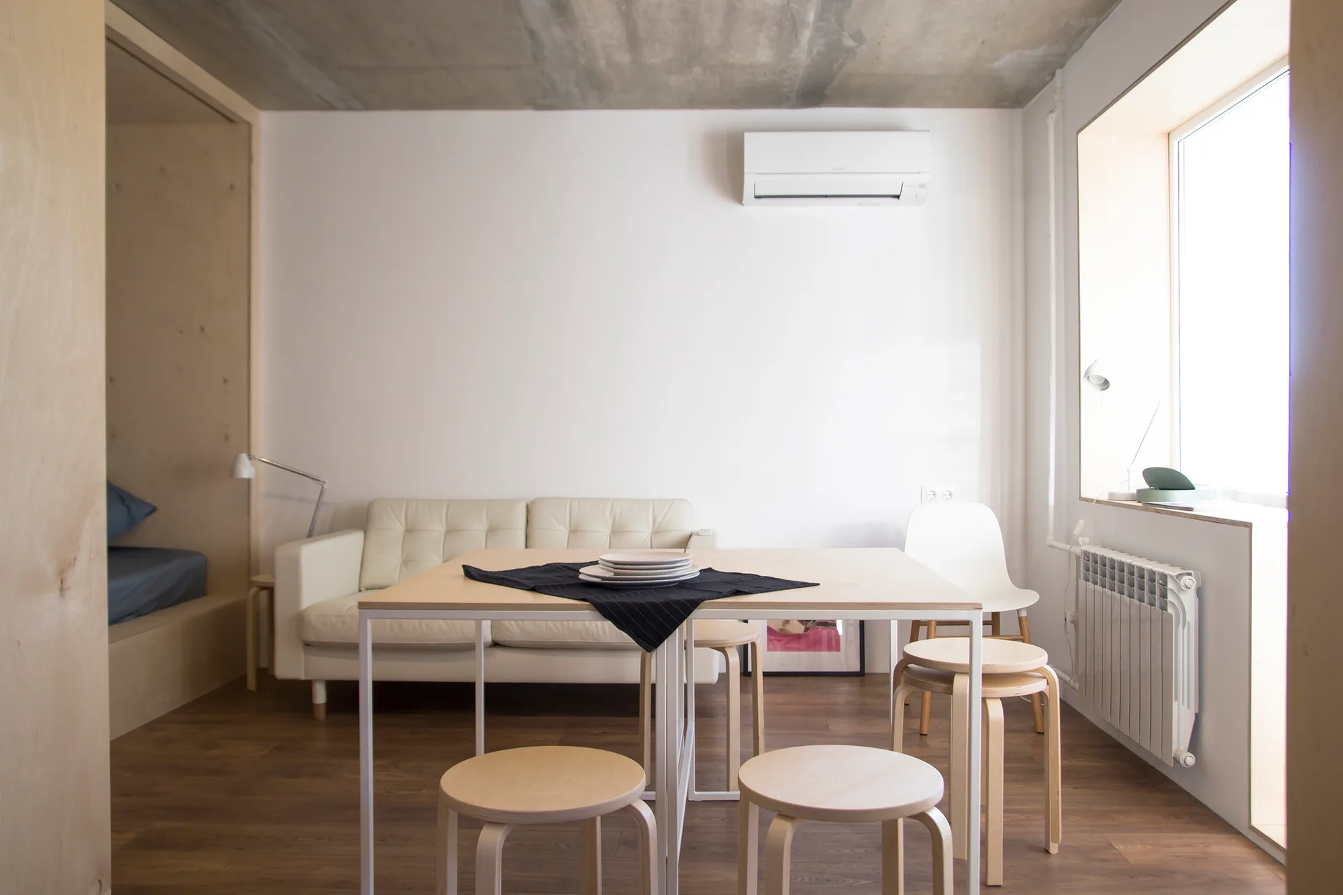 A light wood dining table with stools stands in a minimalist room with a cream sofa, concrete ceiling, and plywood-lined sleeping nook.