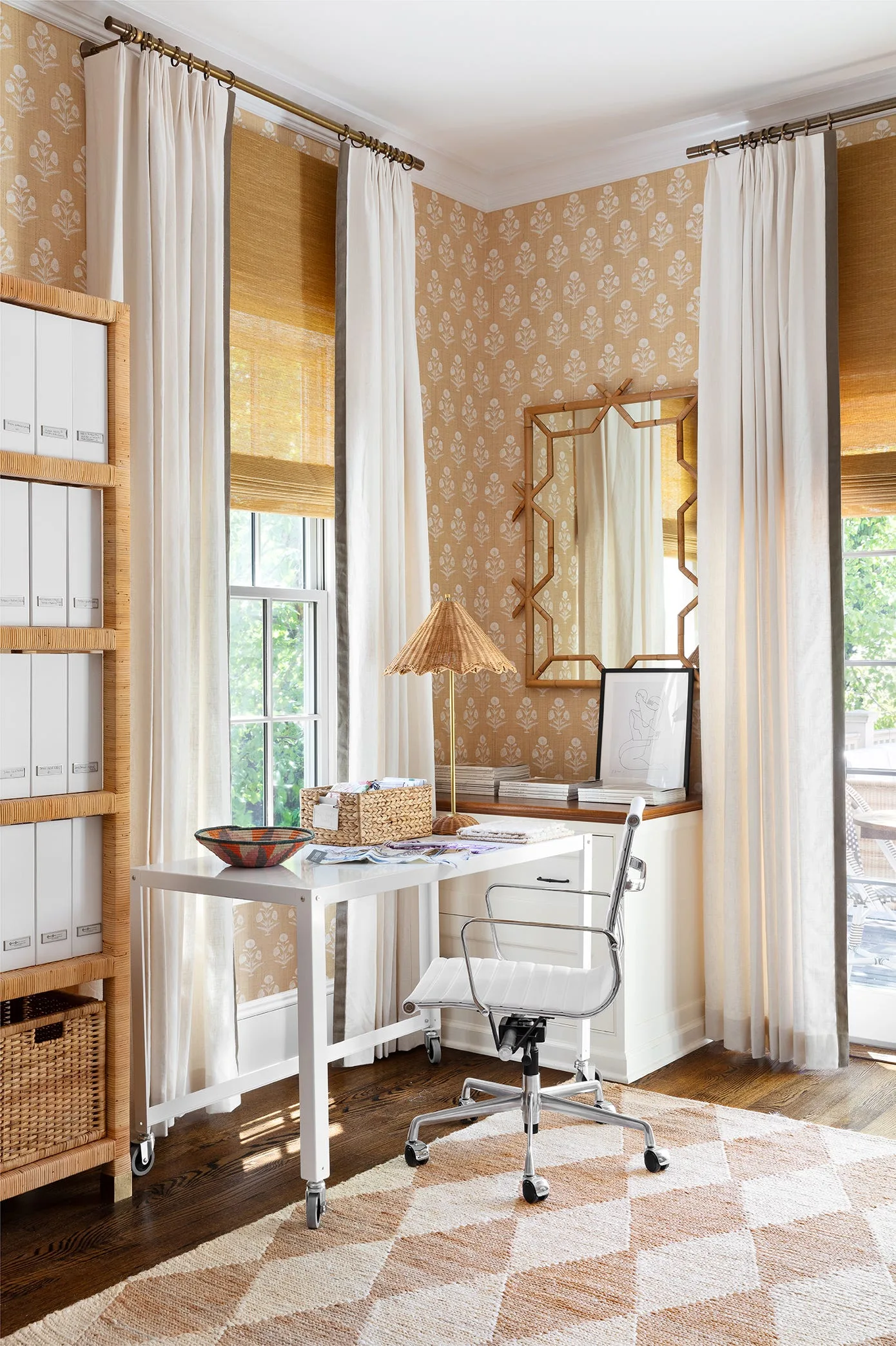 A bright corner home office features a white desk, a white swivel chair, and botanical wallpaper next to windows with woven wood shades.