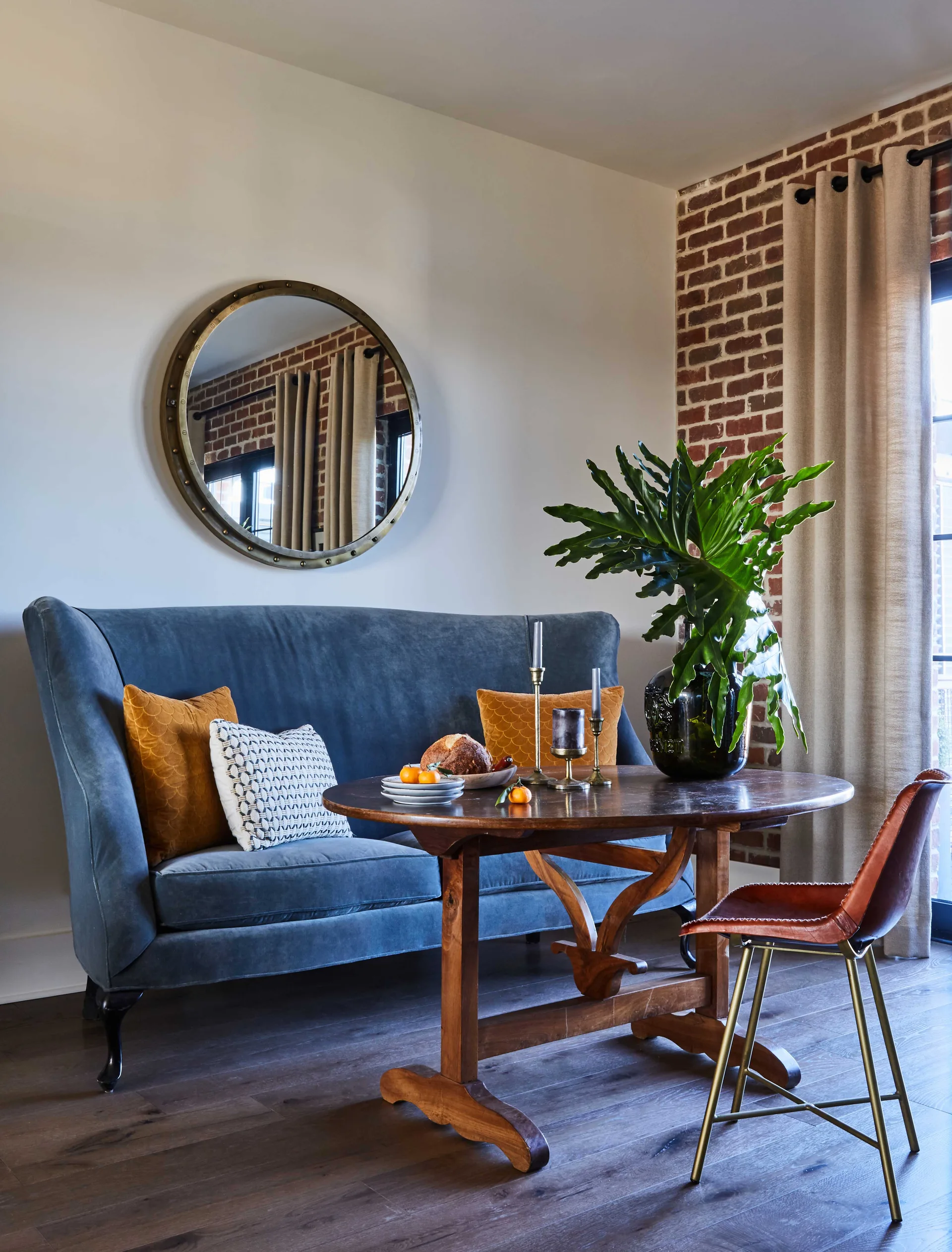 A blue velvet high-back sofa serves as dining seating at a round wooden table next to an exposed brick wall and a round brass mirror.
