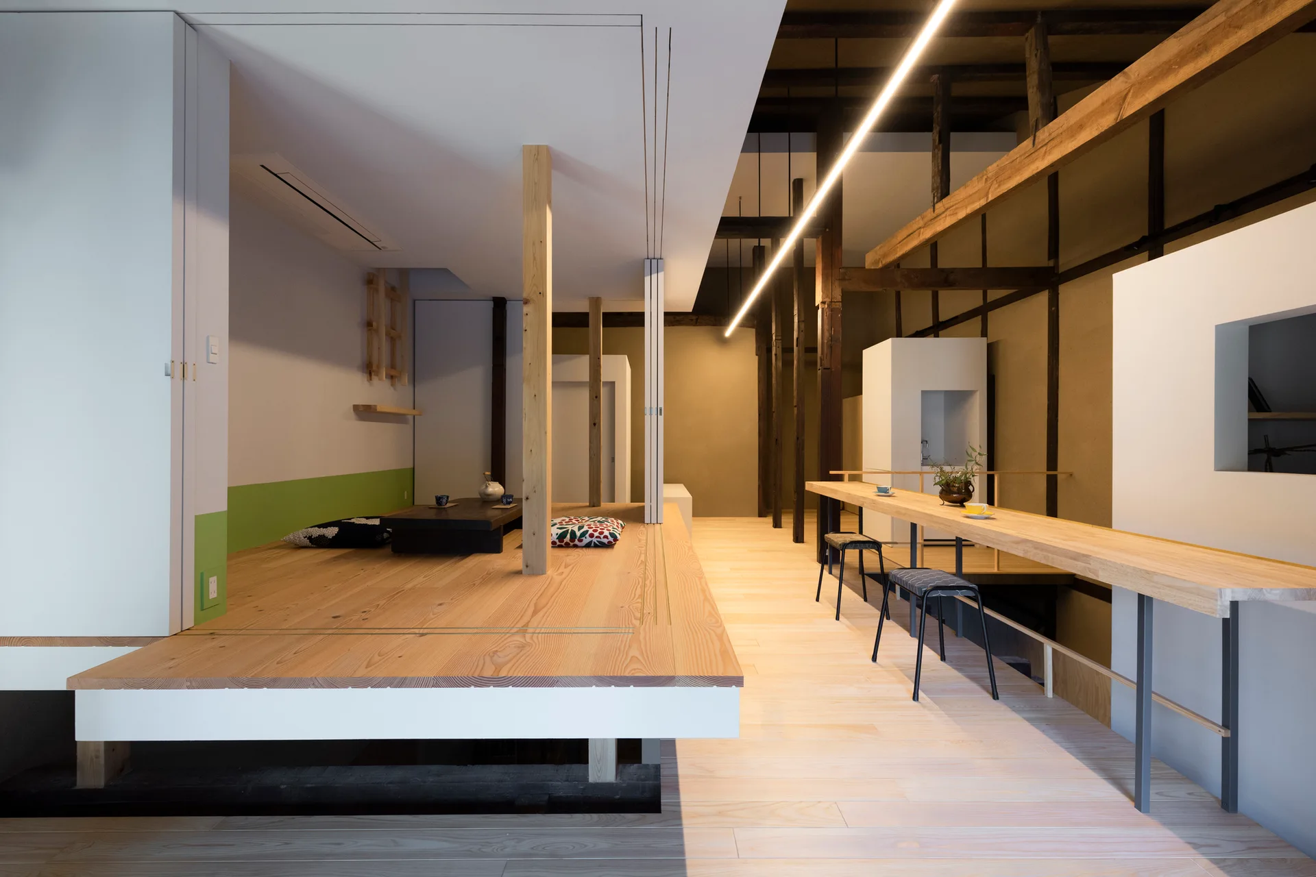A modern Japanese minimalist interior features a raised light-wood platform with cushions next to a long wooden table with black stools.
