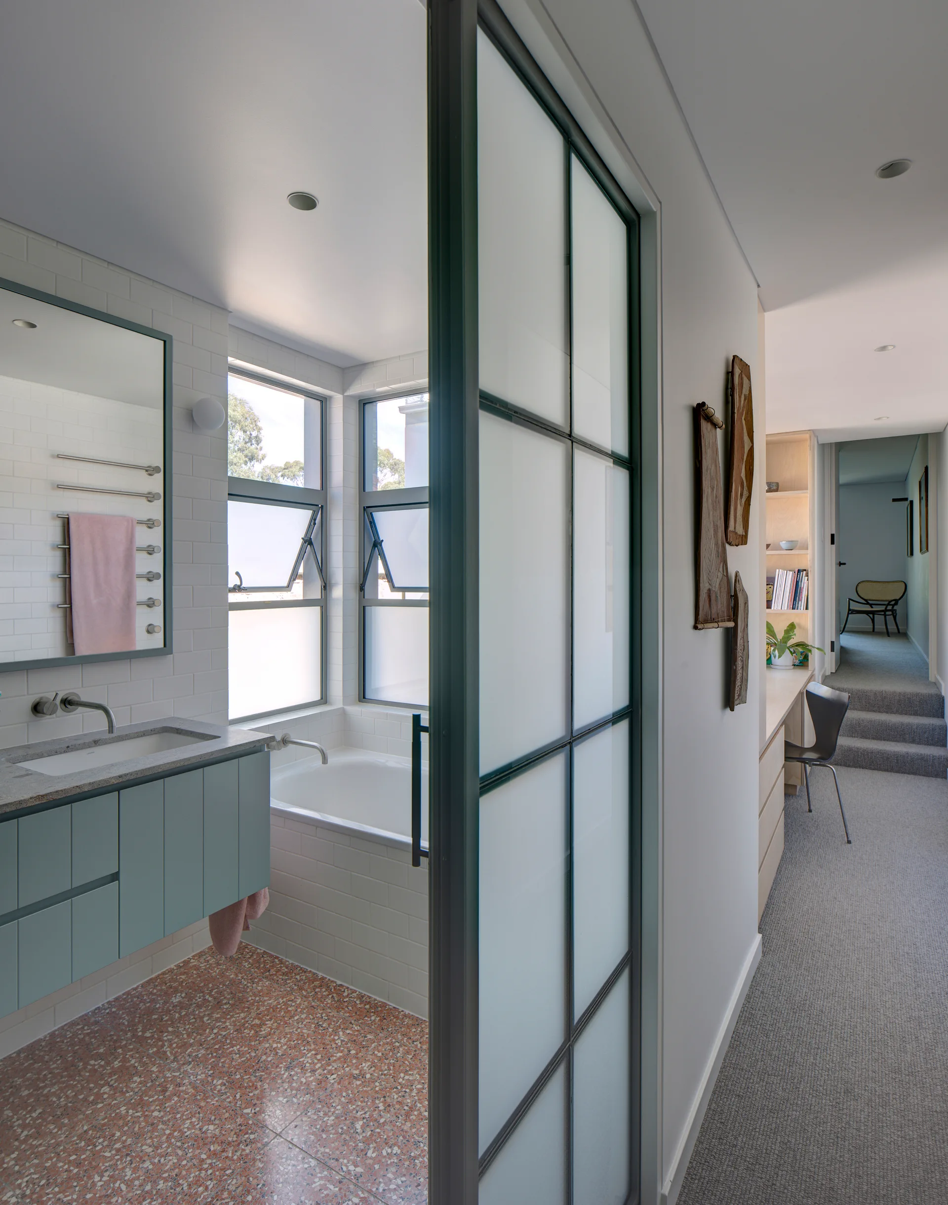 A modern bathroom with a blue-green floating vanity, pink terrazzo flooring, a built-in tub, and a metal-framed frosted glass door.