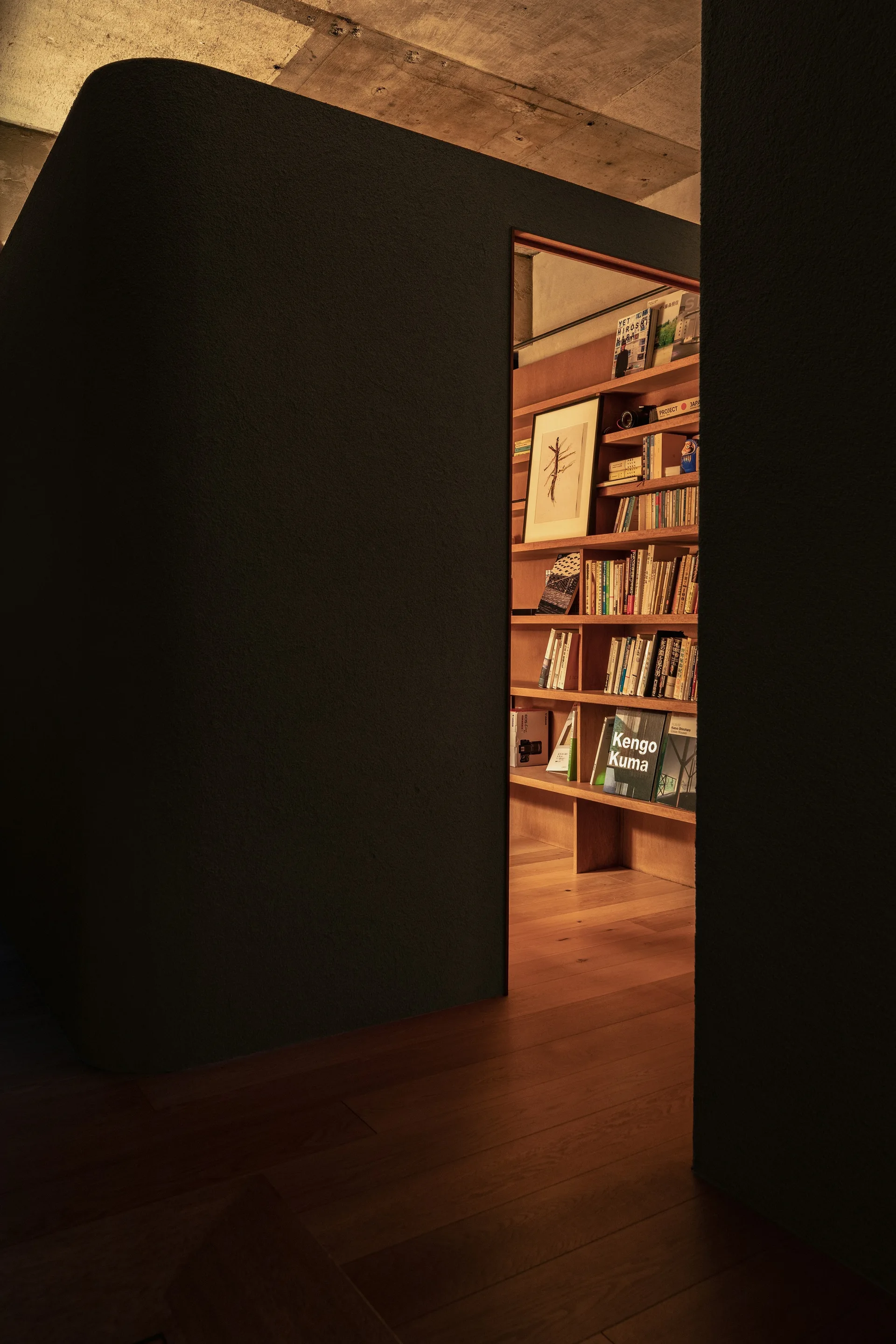 A doorway in a curved charcoal wall opens to a warmly lit study with wooden bookshelves, wood flooring, and an exposed concrete ceiling.