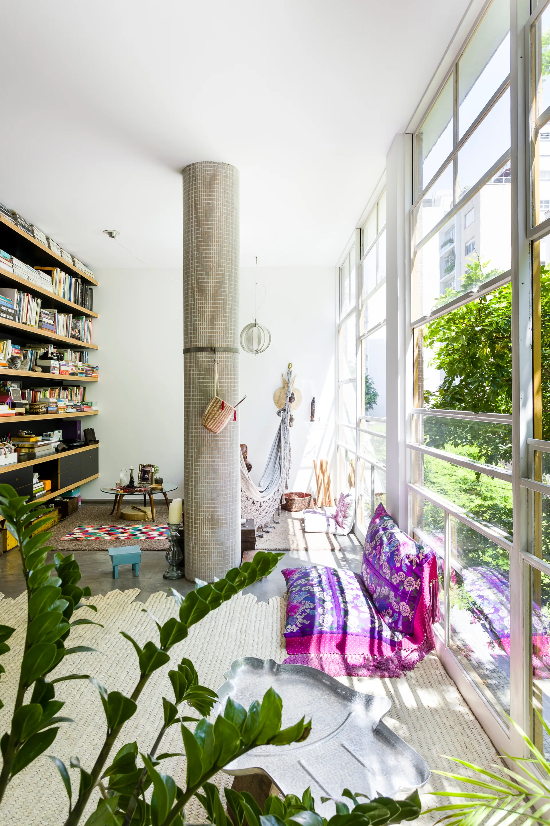 A sunlit bohemian living room features a mosaic tiled structural column next to large grid windows and a purple patterned floor cushion on a woven rug.