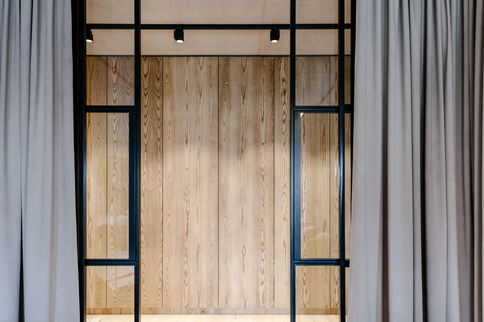 A vertical light wood plank feature wall framed by black metal glass partitions and beige curtains hanging on both sides.