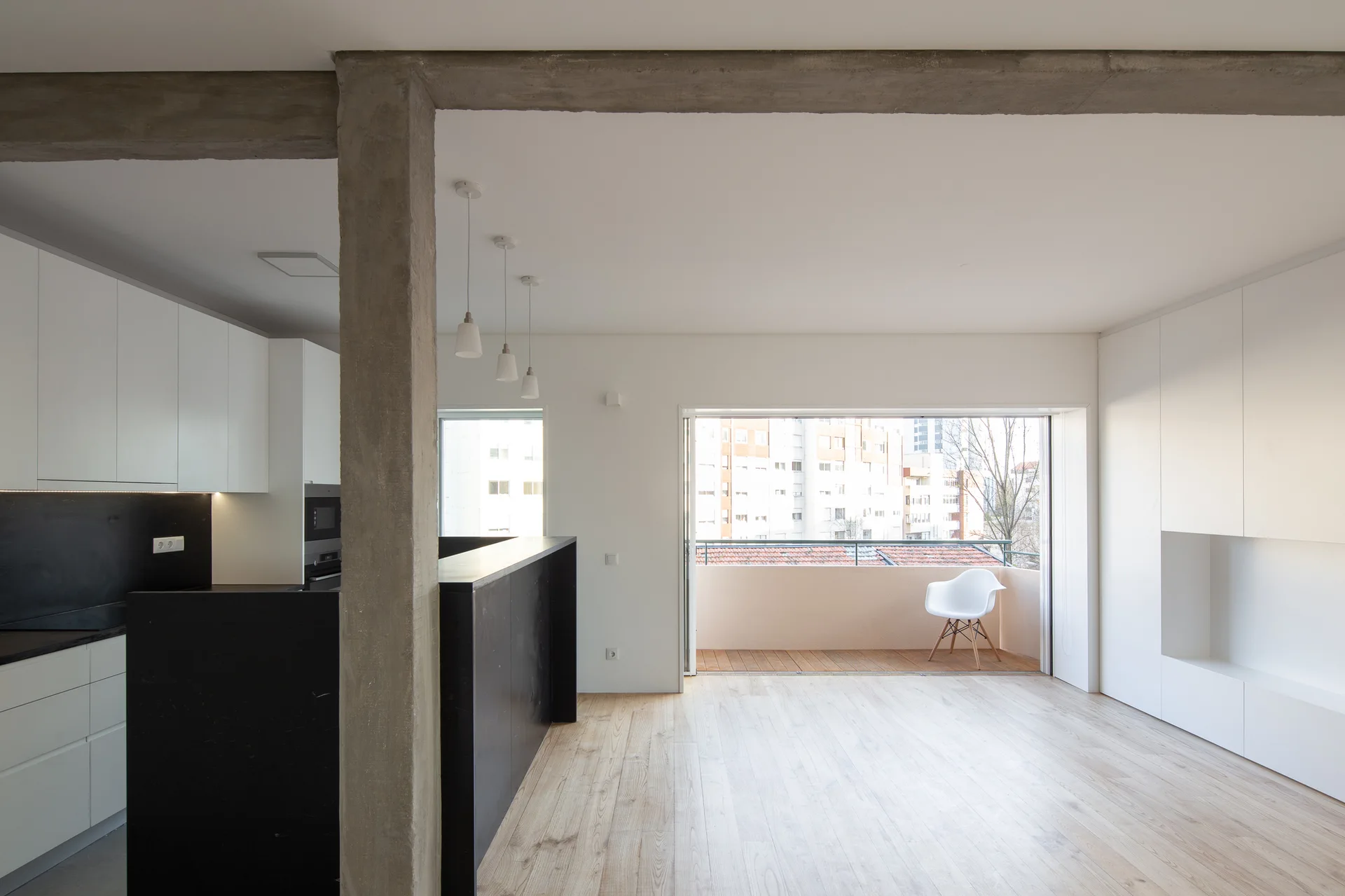 An open-concept minimalist kitchen featuring white cabinets, a stark black island, light wood flooring, and an exposed raw concrete ceiling beam.
