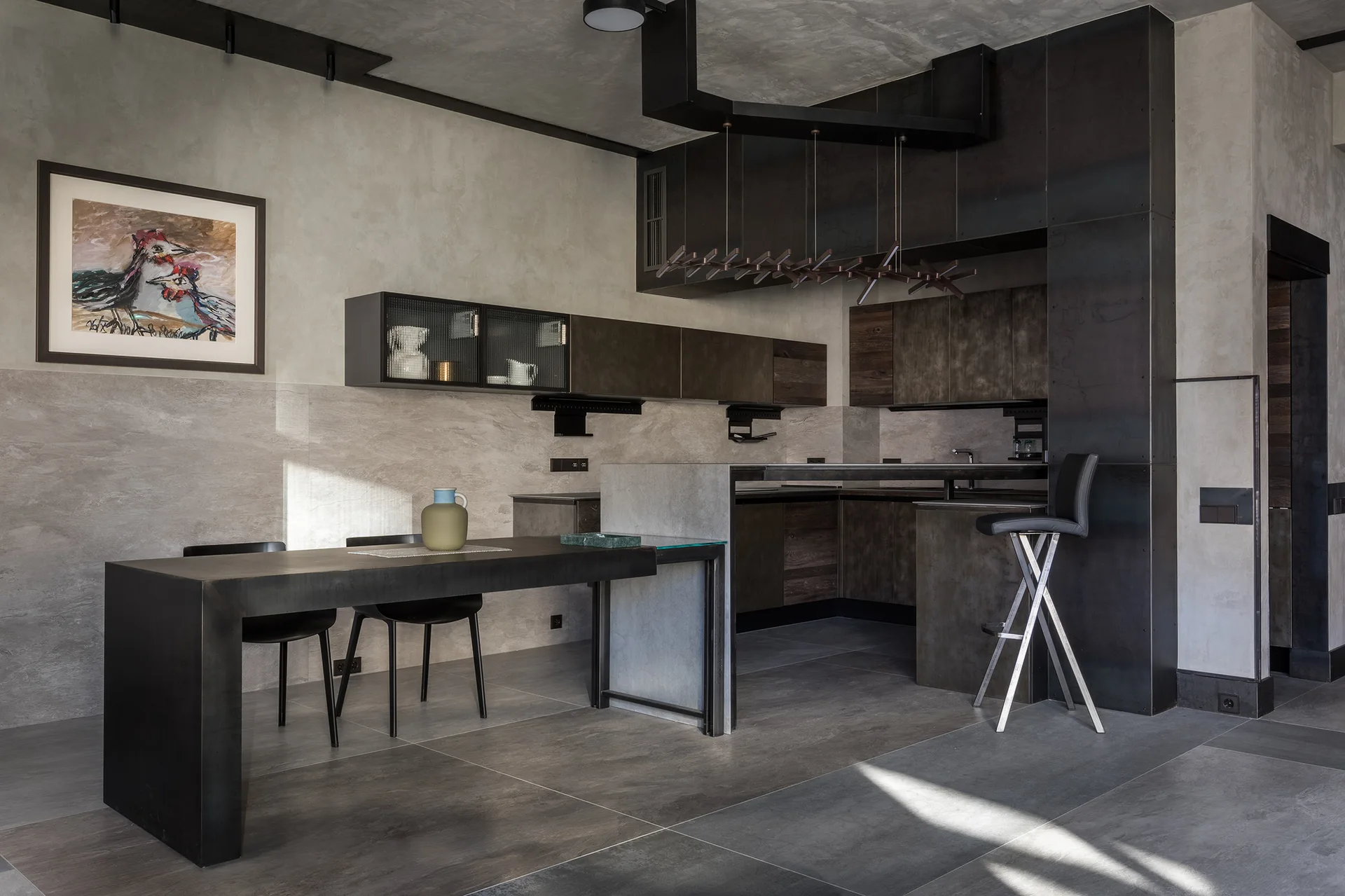 A modern industrial kitchen features dark metal cabinetry, an integrated dining table with black chairs, and concrete-finish walls and floors.