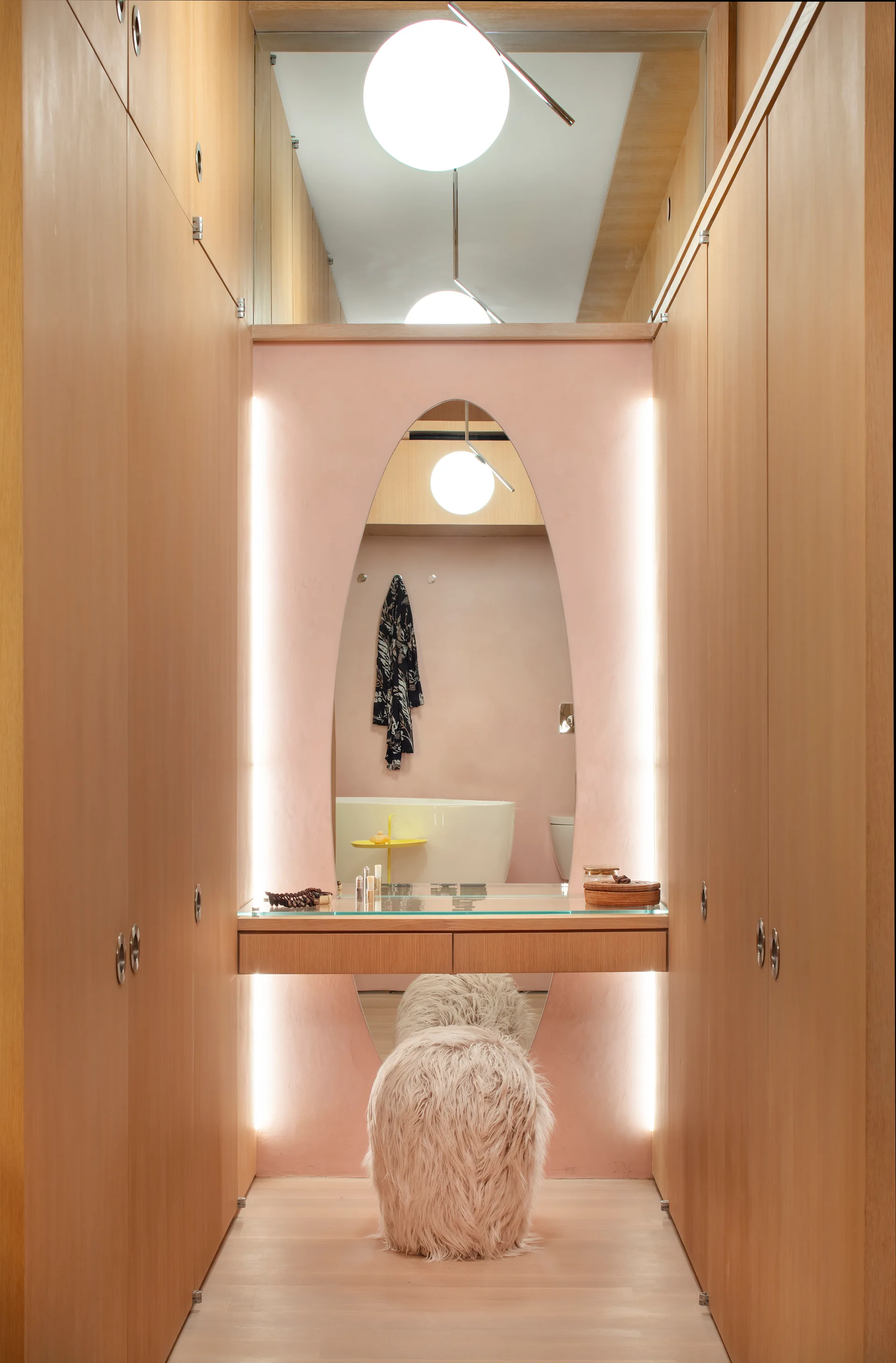 A narrow dressing room features light wood closets, a floating wood vanity with an oval mirror on a pale pink wall, and a furry stool.
