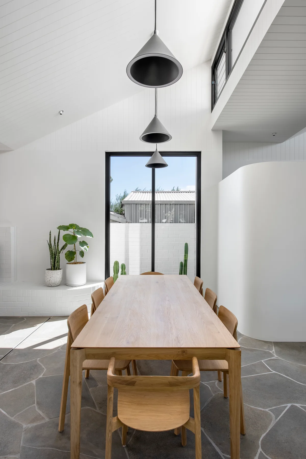 A light wood dining table and chairs are set on grey flagstone flooring beneath a vaulted white ceiling and three grey cone pendant lights.
