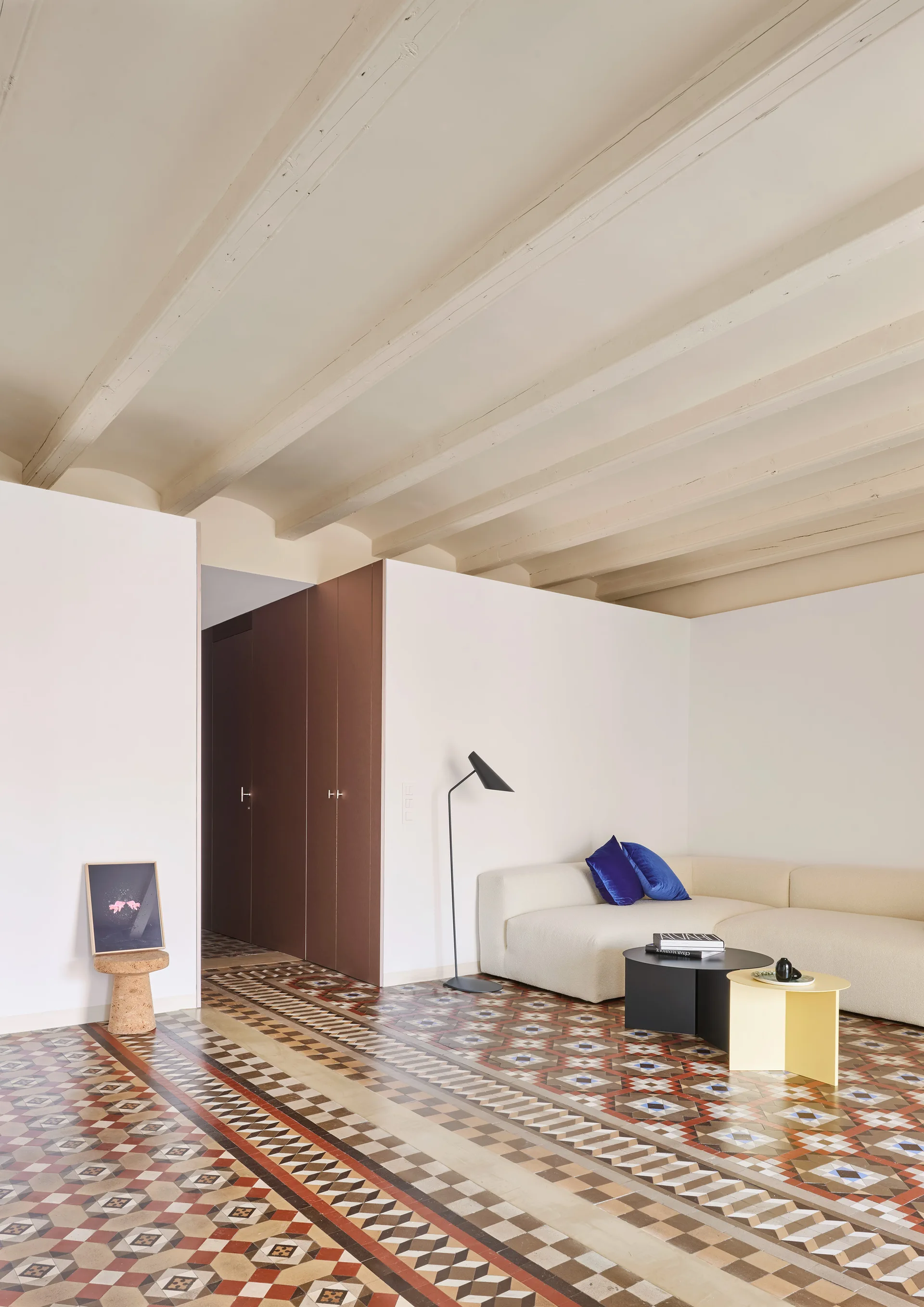 A contemporary living room featuring a cream modular sofa, nesting coffee tables, exposed ceiling beams, and a colorful geometric tiled floor.