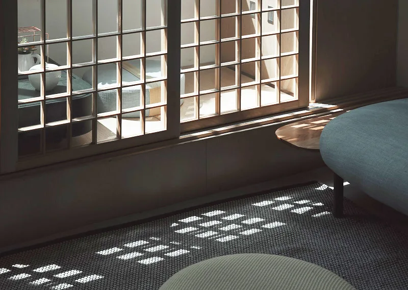 A wood lattice sliding screen door casts grid pattern shadows across a textured grey rug and minimalist furniture in a sunlit room.