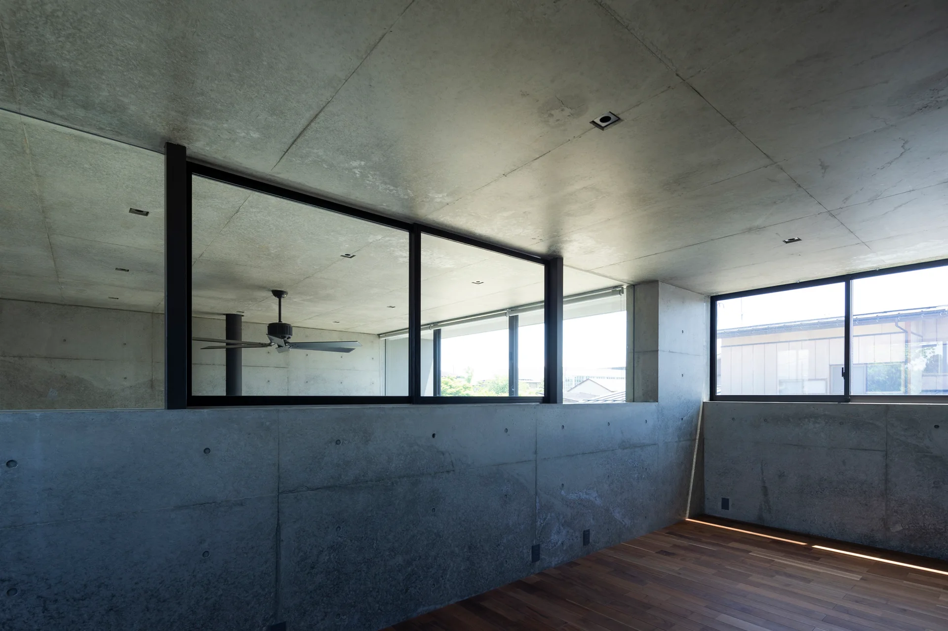 A minimalist room features exposed concrete walls and ceiling, a glass partition window looking into an adjacent space, and dark wood plank flooring.