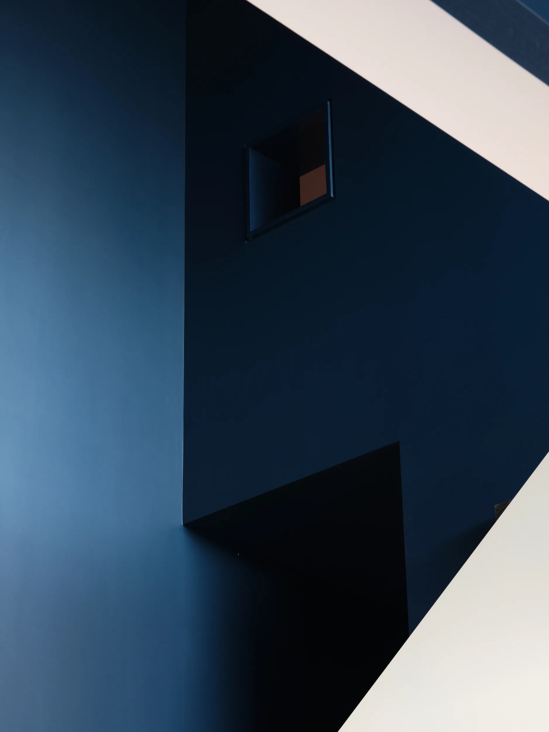 A sharp geometric staircase interior featuring matte navy blue walls, a small square cutout window, and a contrasting white surface.