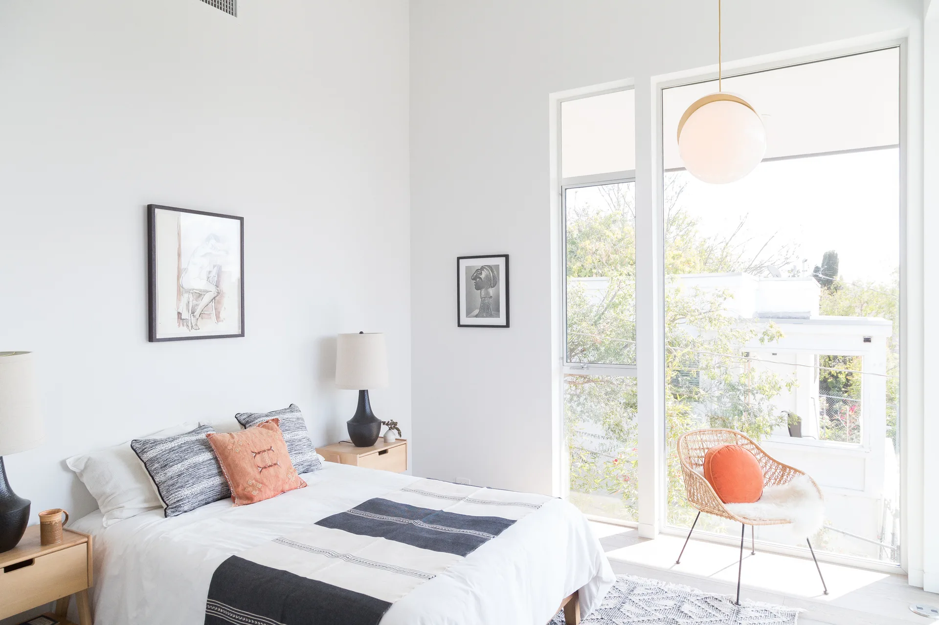 A modern bedroom features white walls, a bed with black and white patterned textiles, light wood nightstands, and a wicker chair near a large window.