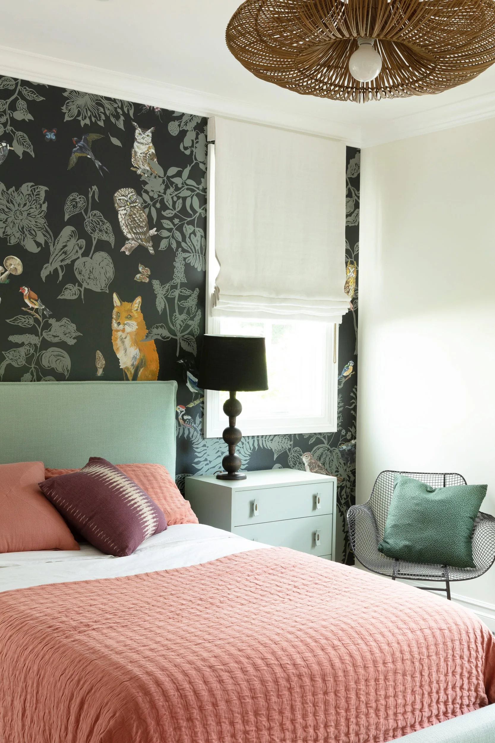 A bedroom featuring a bed with a sage green headboard, pink bedding, a dark woodland animal wallpaper wall, and a black wire chair.