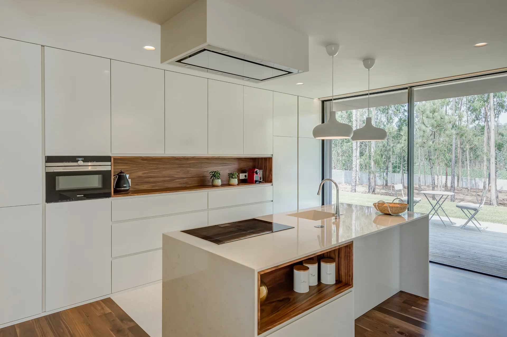 A modern white minimalist kitchen featuring handleless cabinetry, a recessed wood-toned shelf, and a central white island under white pendant lights.